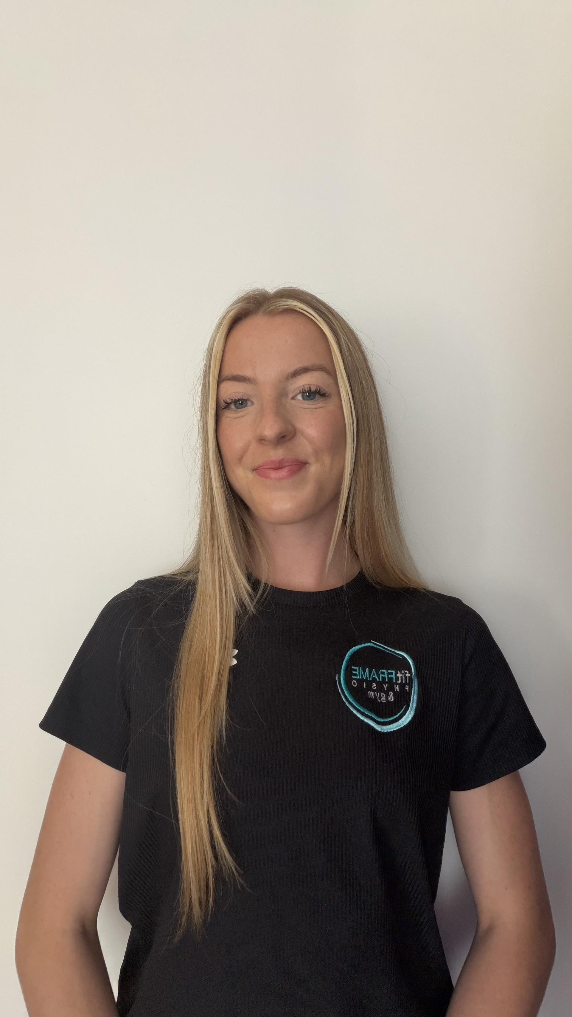 A woman with long blonde hair wearing a black shirt with a logo that reads 'FIT FRAME FITNESS & GYM' stands against a plain white wall.