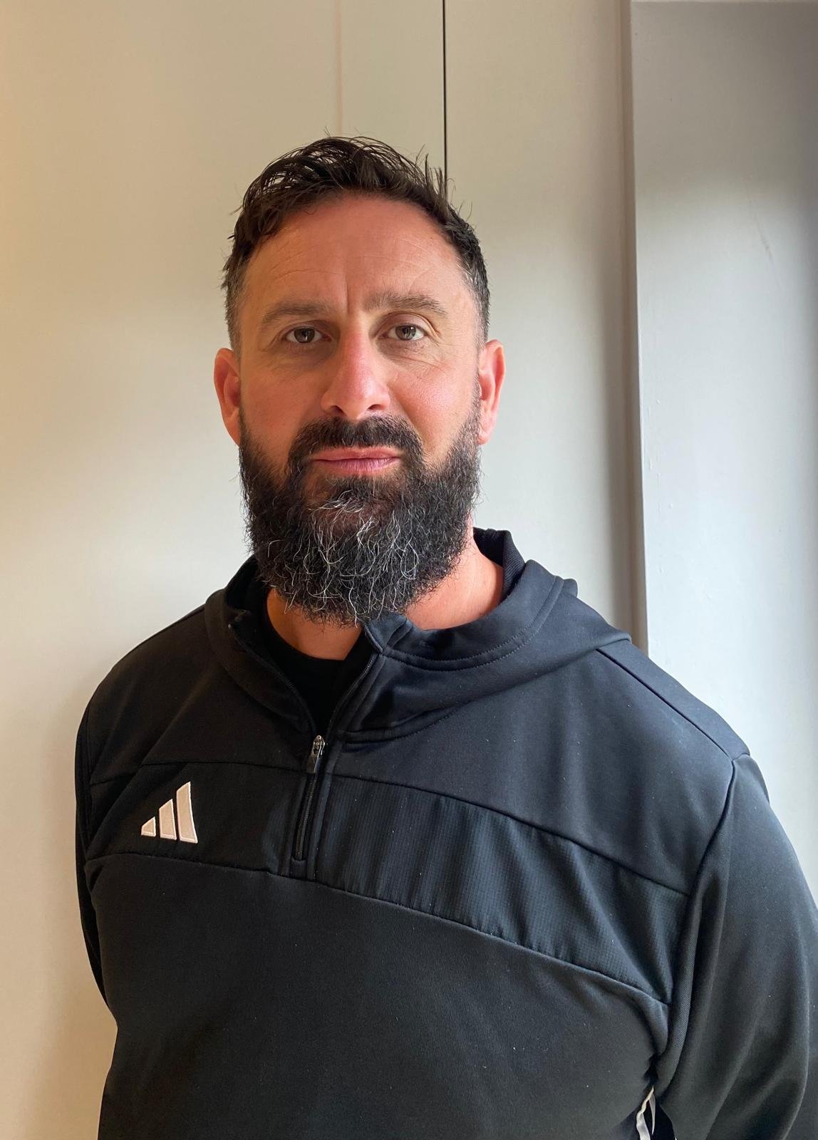 A man with dark hair, a beard, and wearing a red and blue plaid shirt in a kitchen. There is a poster on the wall with a figure holding a pizza slice.