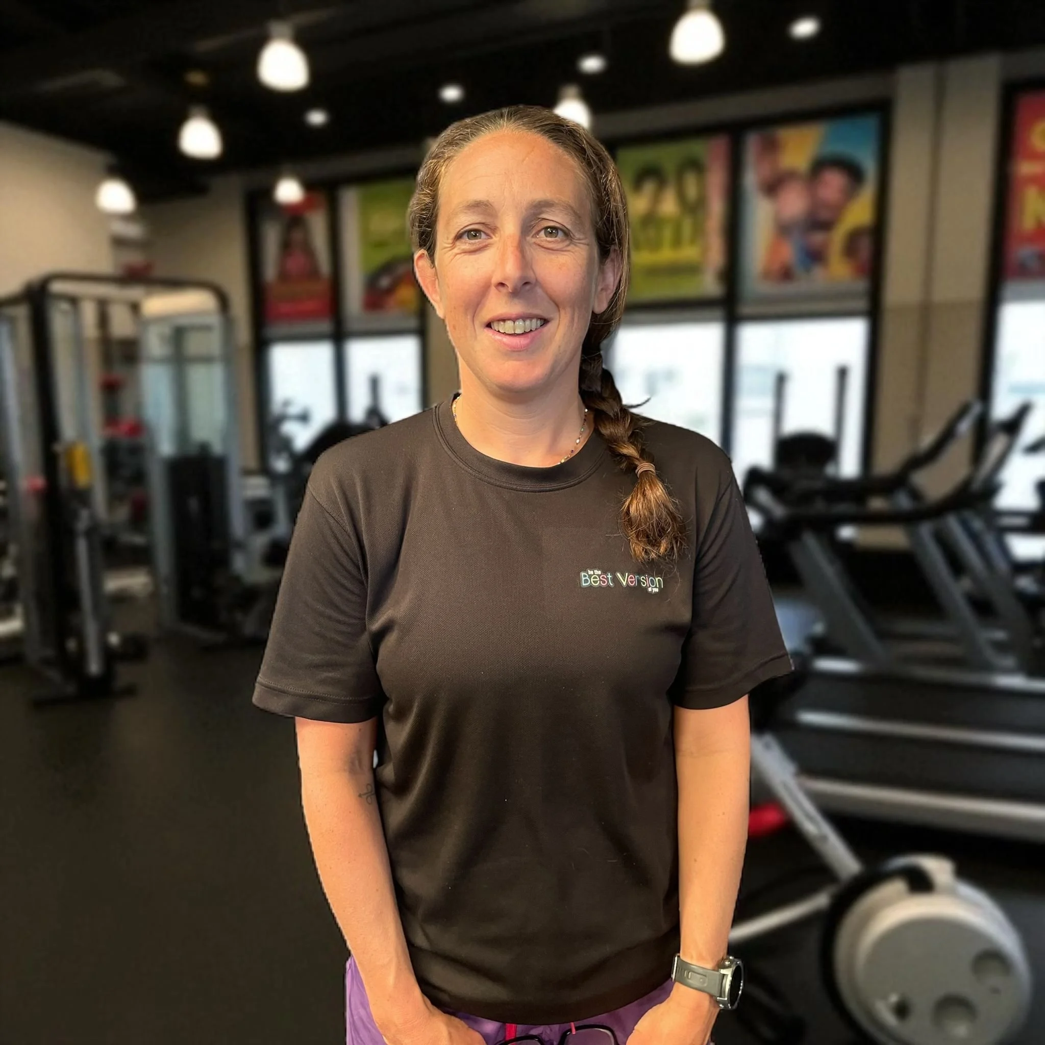 A woman with brown hair in a braid wearing a black "Best Version" t-shirt standing in a gym with exercise machines and colorful posters in the background.