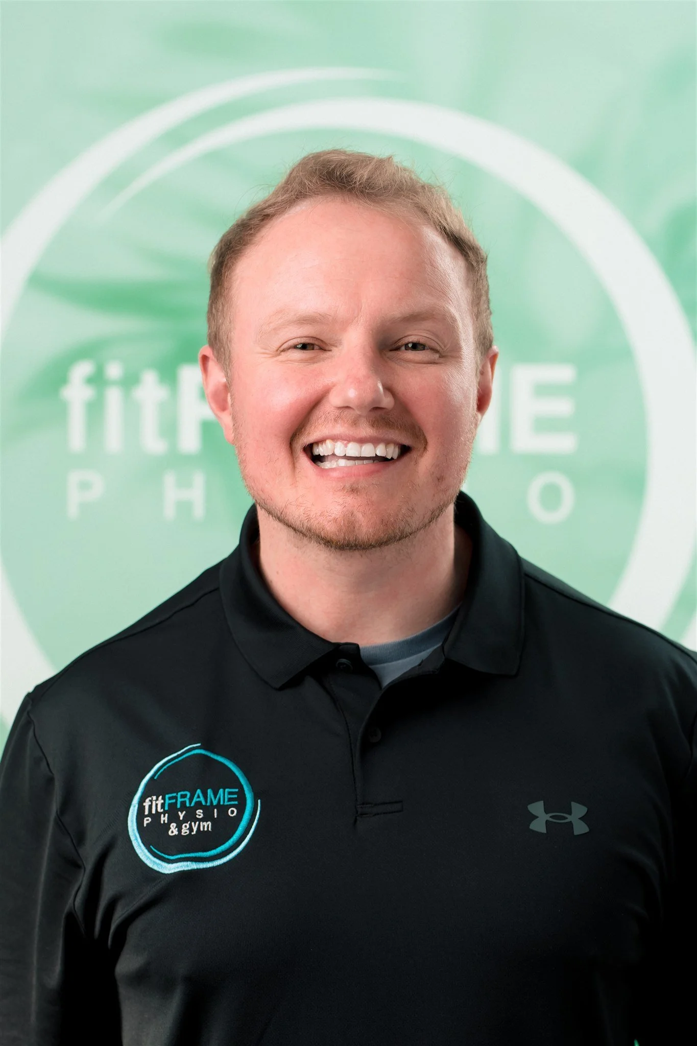 A smiling young man with light-colored hair, wearing a dark Under Armour t-shirt, standing against a plain white wall.