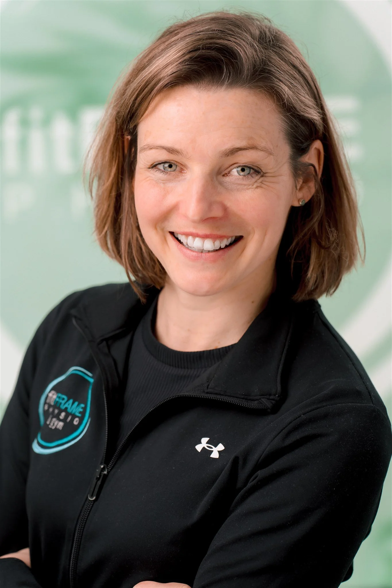 A woman with short brown hair and blue eyes smiling, wearing a black jacket with an Under Armour logo and a patch that says 'FRAME