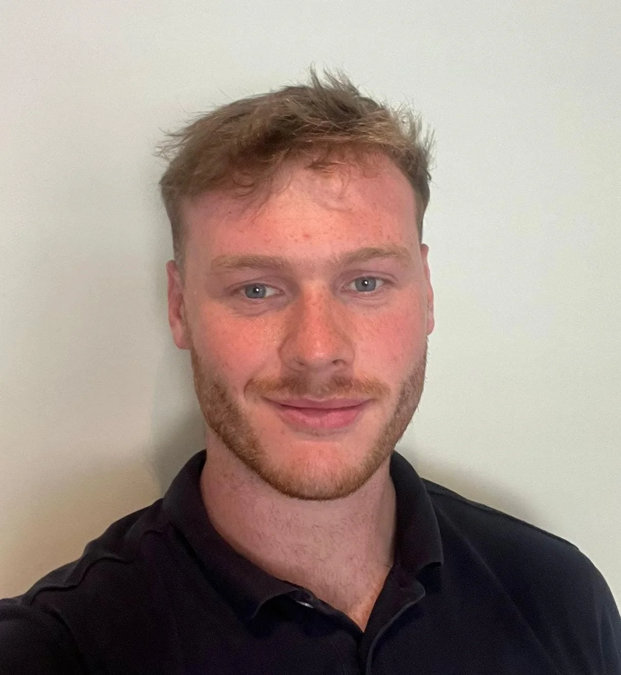 A young man with light skin, red hair, and a beard, wearing a black shirt, smiling against a plain light-colored wall.