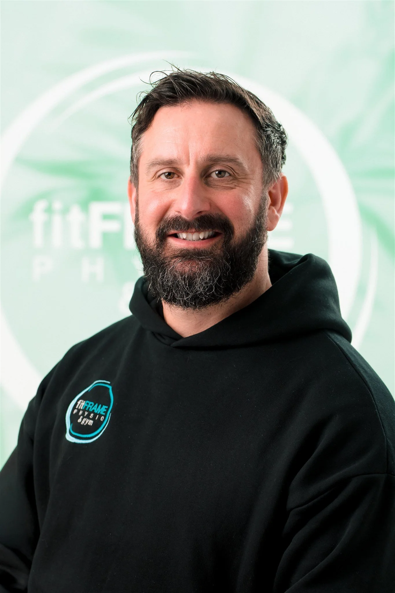 A man with dark hair, a beard, and wearing a red and blue plaid shirt in a kitchen. There is a poster on the wall with a figure holding a pizza slice.