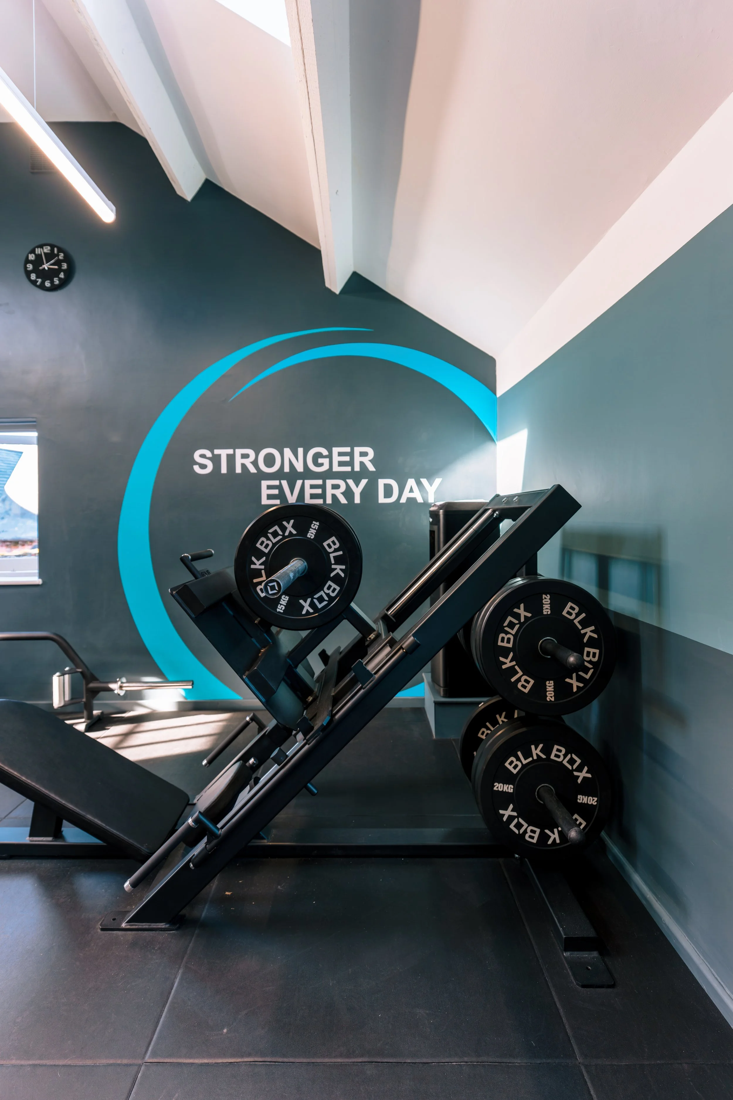 A gym with an inclined weightlifting machine and several weight plates against a teal wall. The wall has a motivational message, 'STRONGER EVERY DAY,' and a clock showing 4:11.