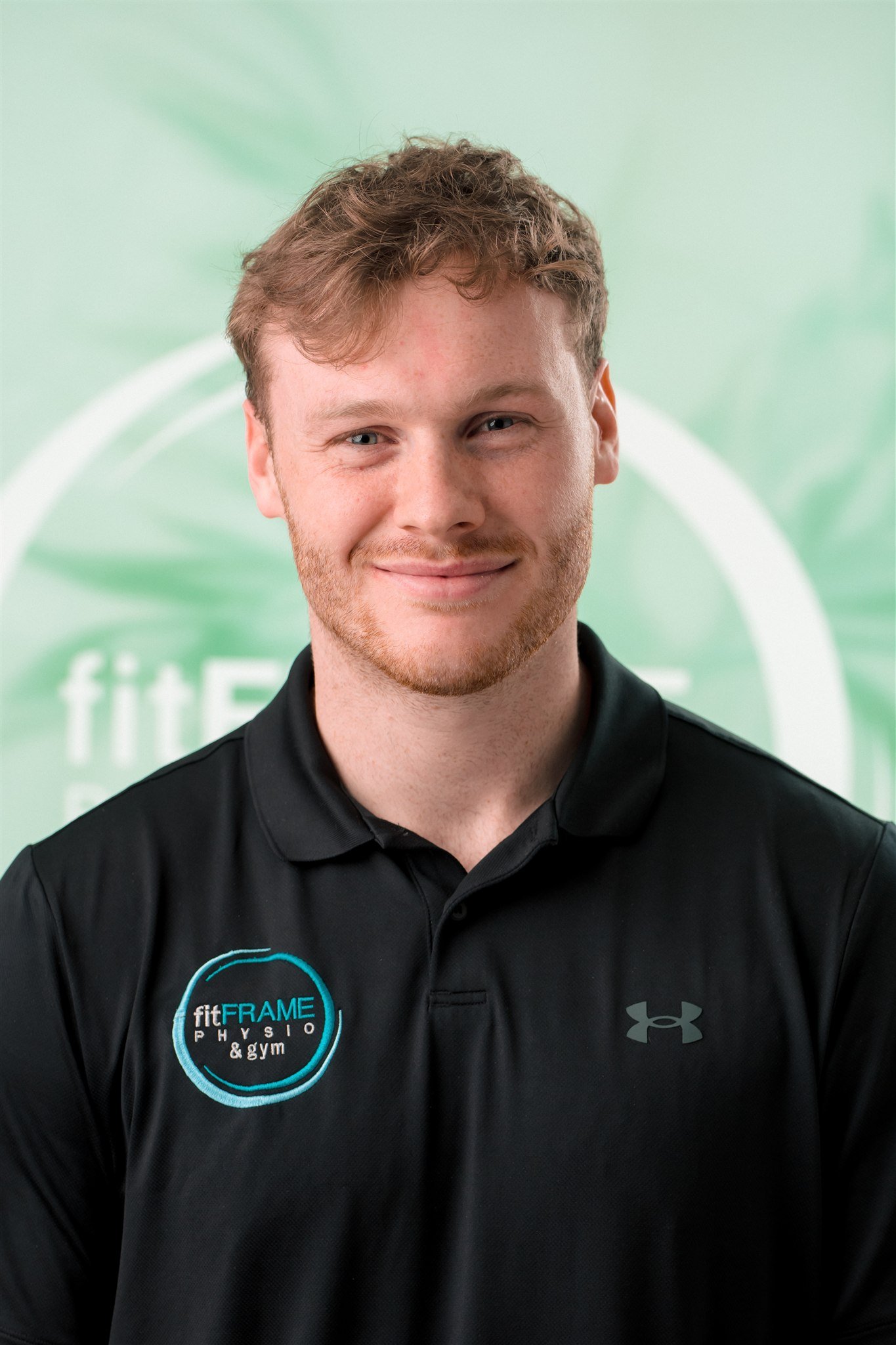A young man with light skin, red hair, and a beard, wearing a black shirt, smiling against a plain light-colored wall.