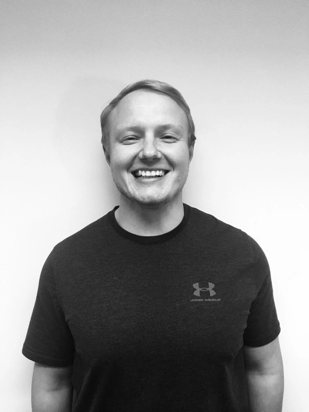 A smiling young man with light-colored hair, wearing a dark Under Armour t-shirt, standing against a plain white wall.