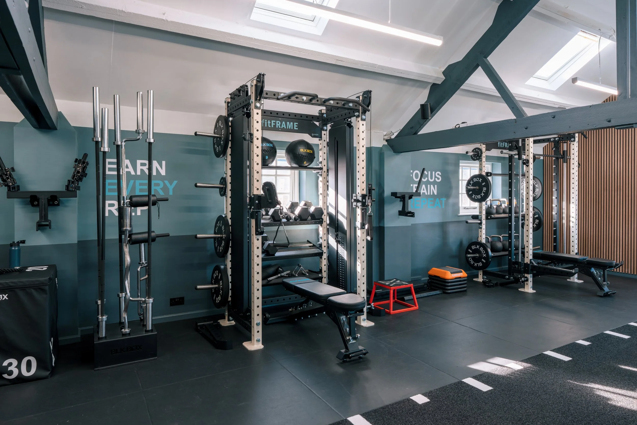 A modern gym with workout equipment including barbells, weight plates, a bench, a squat rack, and a small red step platform, all set against a blue and white wall with motivational phrases.