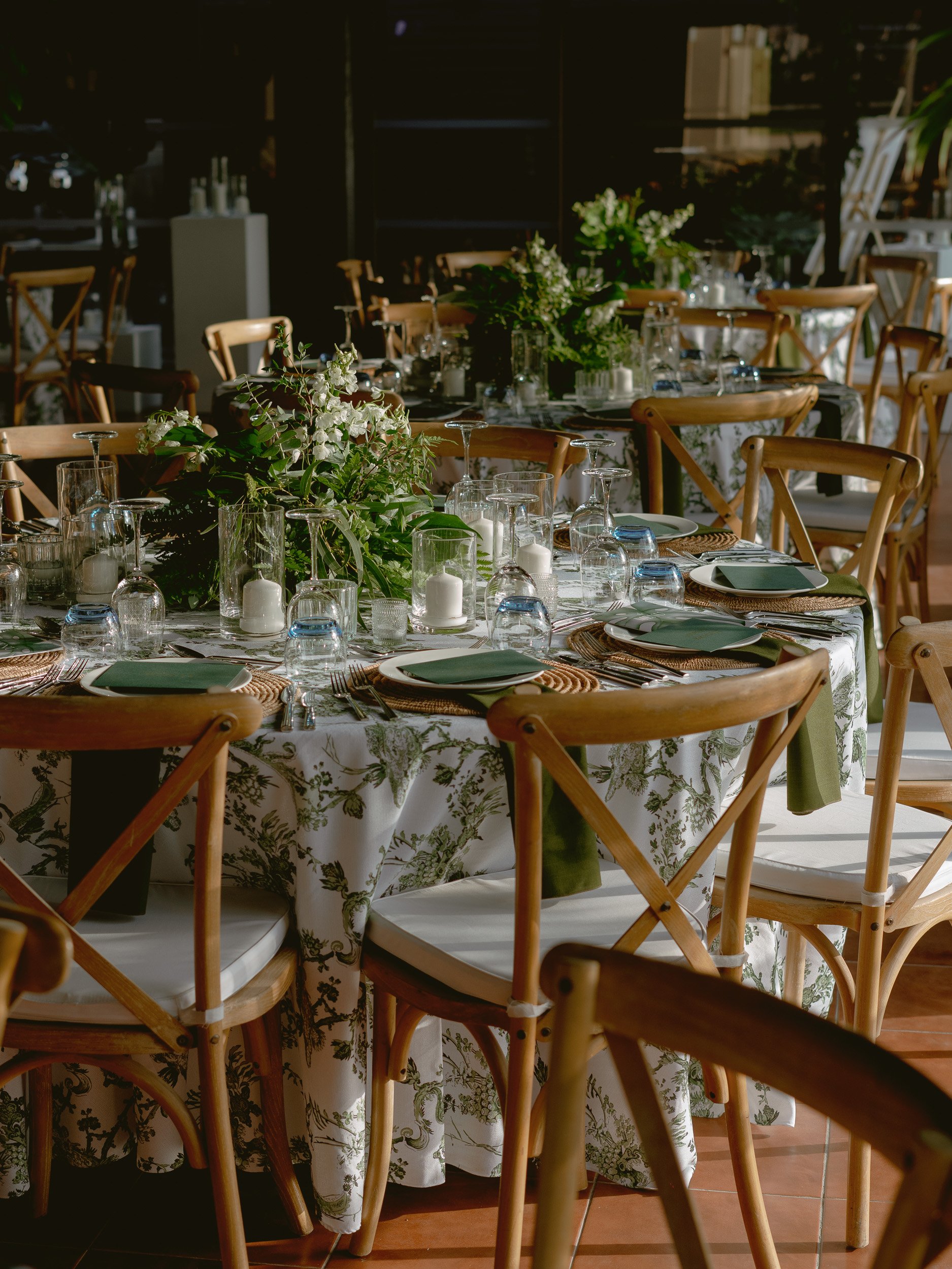 A round banquet table set for a special event, decorated with a floral centerpiece, candles, and tableware, in a well-lit room with dark background and wooden chairs.