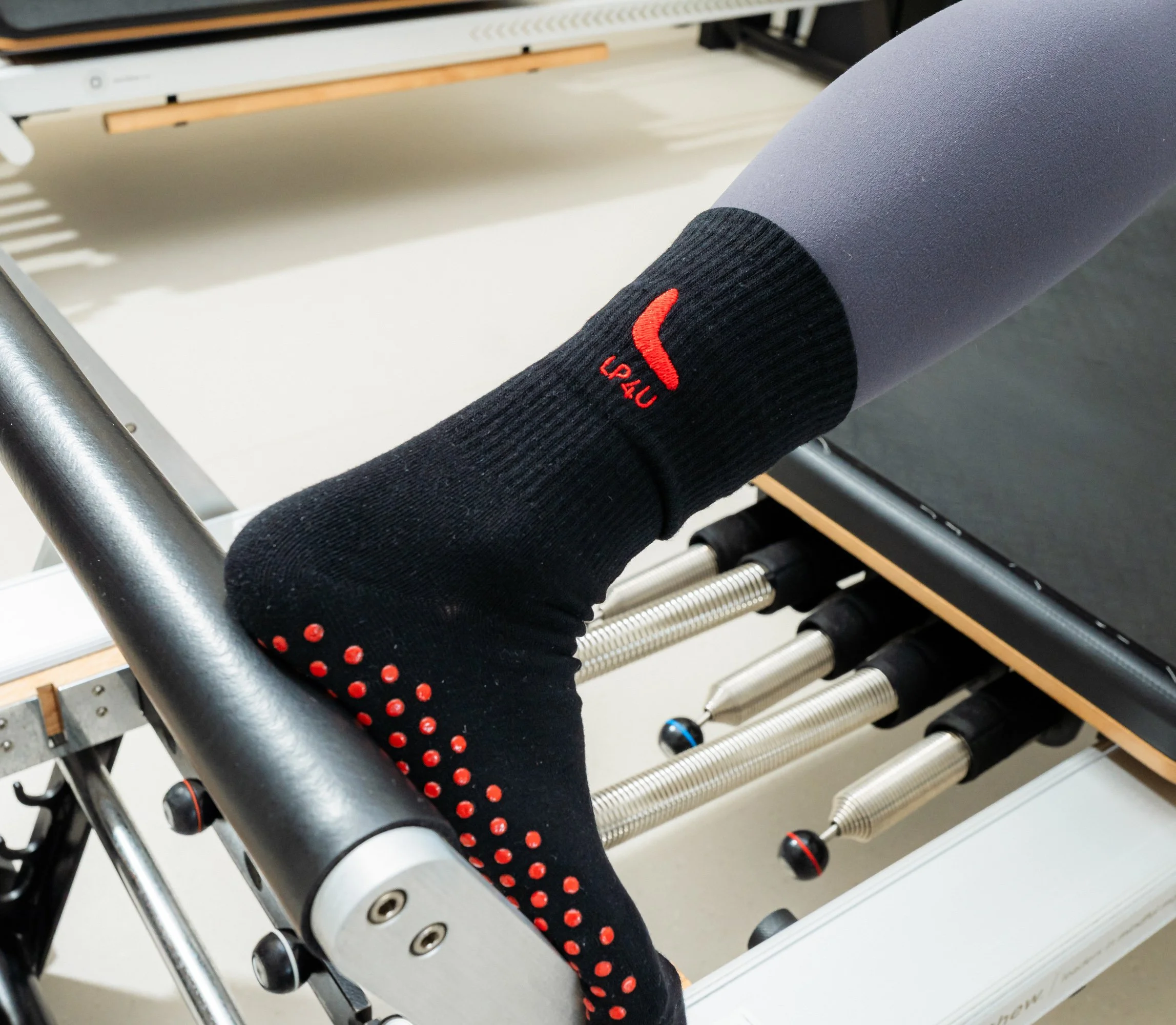 Close-up of a person's leg wearing black athletic compression socks with red dots and the brand logo 'LULU' in red, resting on a Pilates reformer machine's footbar in a fitness studio.