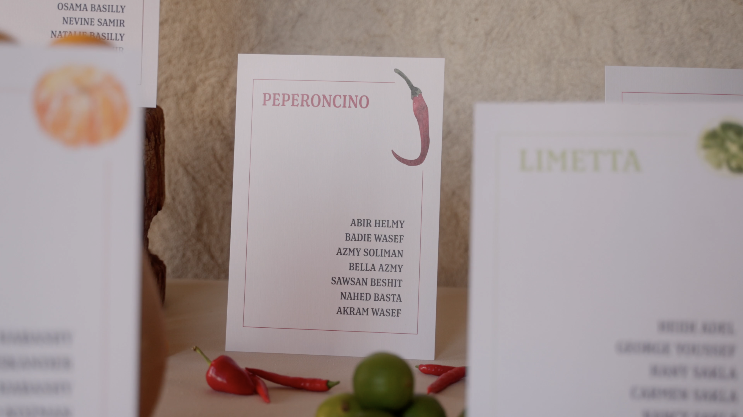 Display of name cards in front of a beige wall, including a card labeled "PEPERONCINO" with a red chili pepper illustration, and other cards for different dishes. There are red and green chili peppers and a small tomato on the surface in front of the