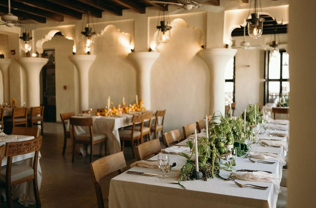Elegant indoor dining area with a long table set for a meal, decorated with green plants and white candles, surrounded by wooden chairs, featuring ornate wall arches and warm lighting.