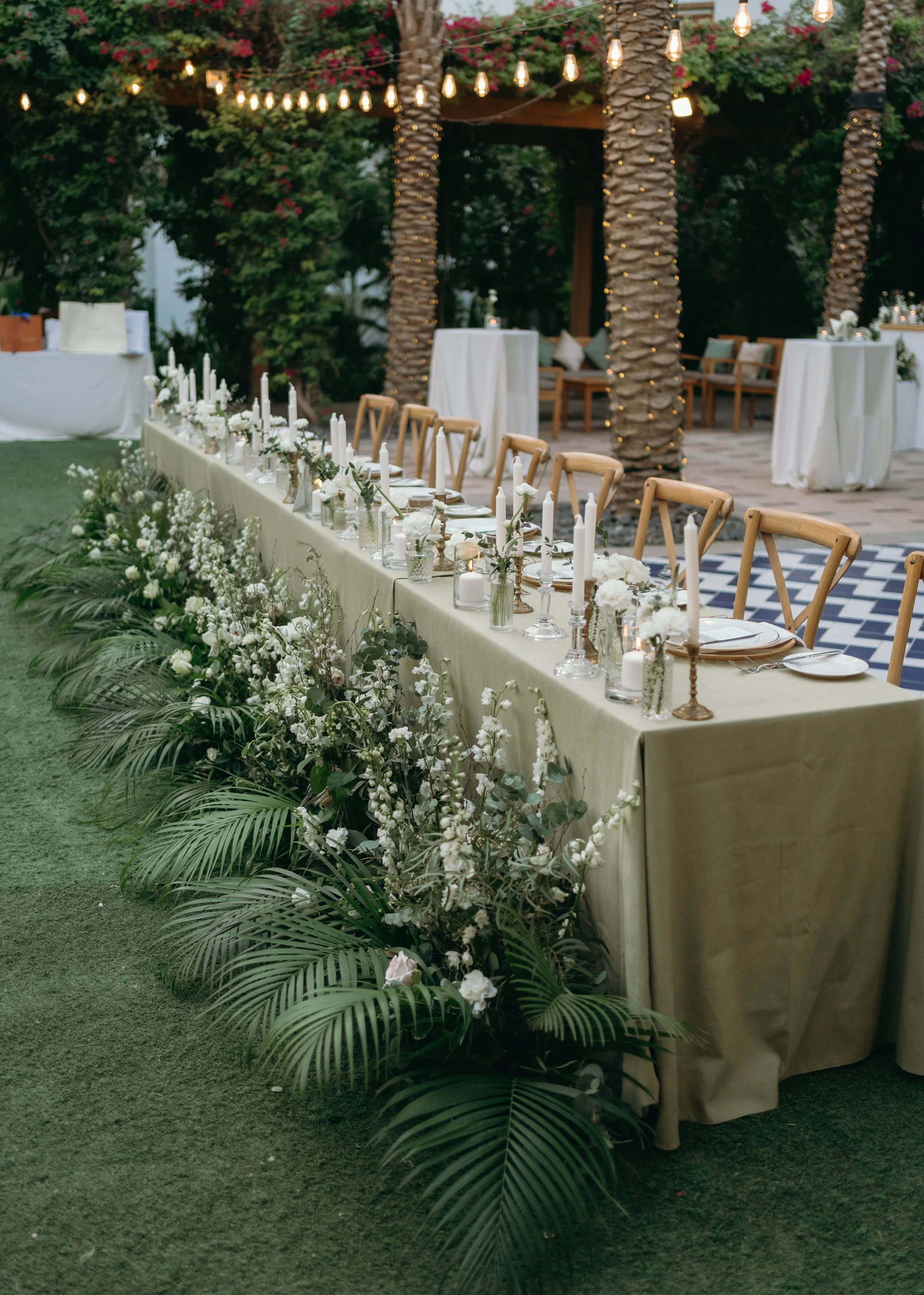 Outdoor event setting with a long banquet table decorated with white flowers, candles, and greenery, surrounded by palm trees and string lights at dusk.