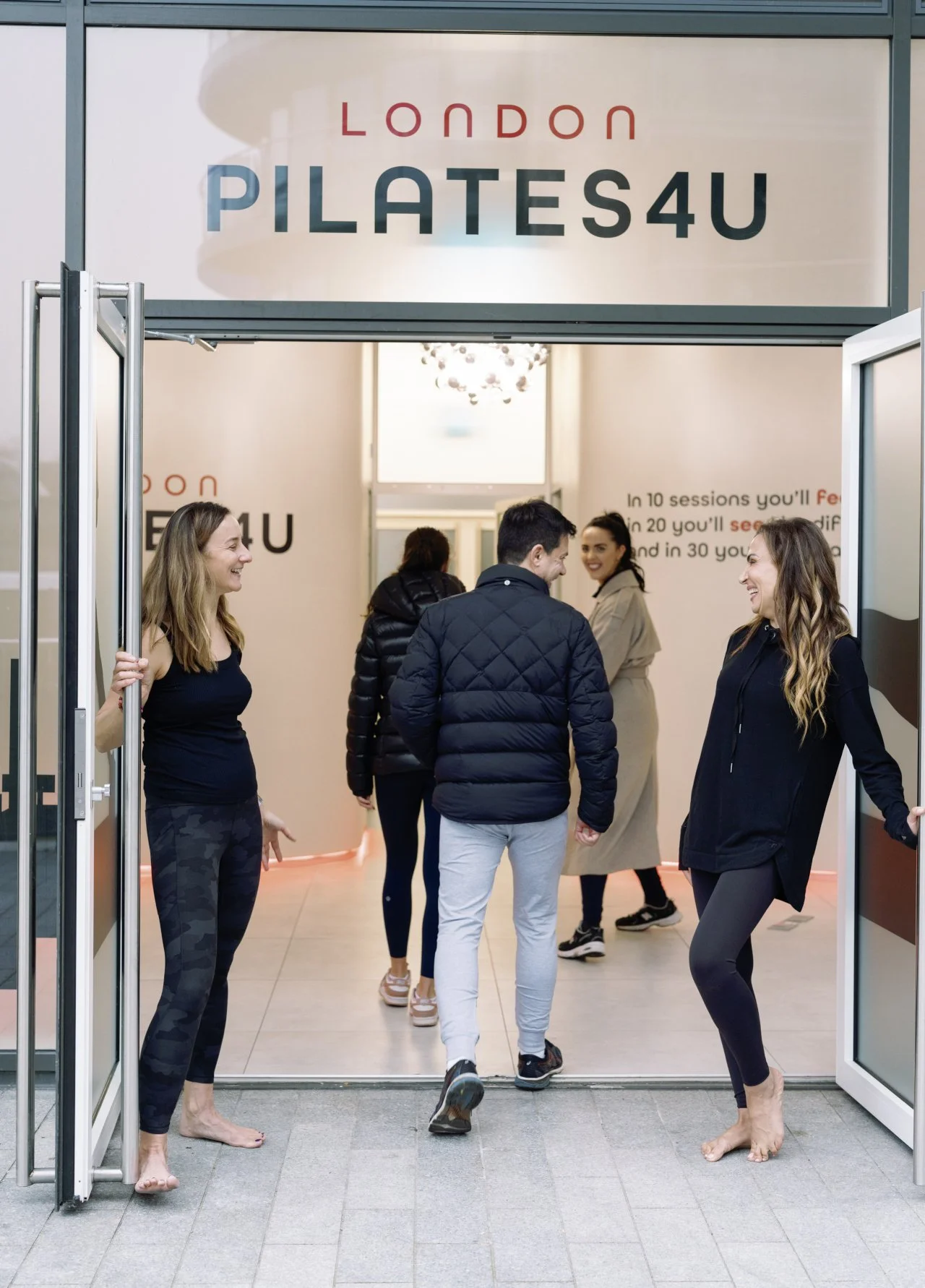 People smiling and chatting at the entrance of a Pilates studio with a sign reading 'London Pilates4U'