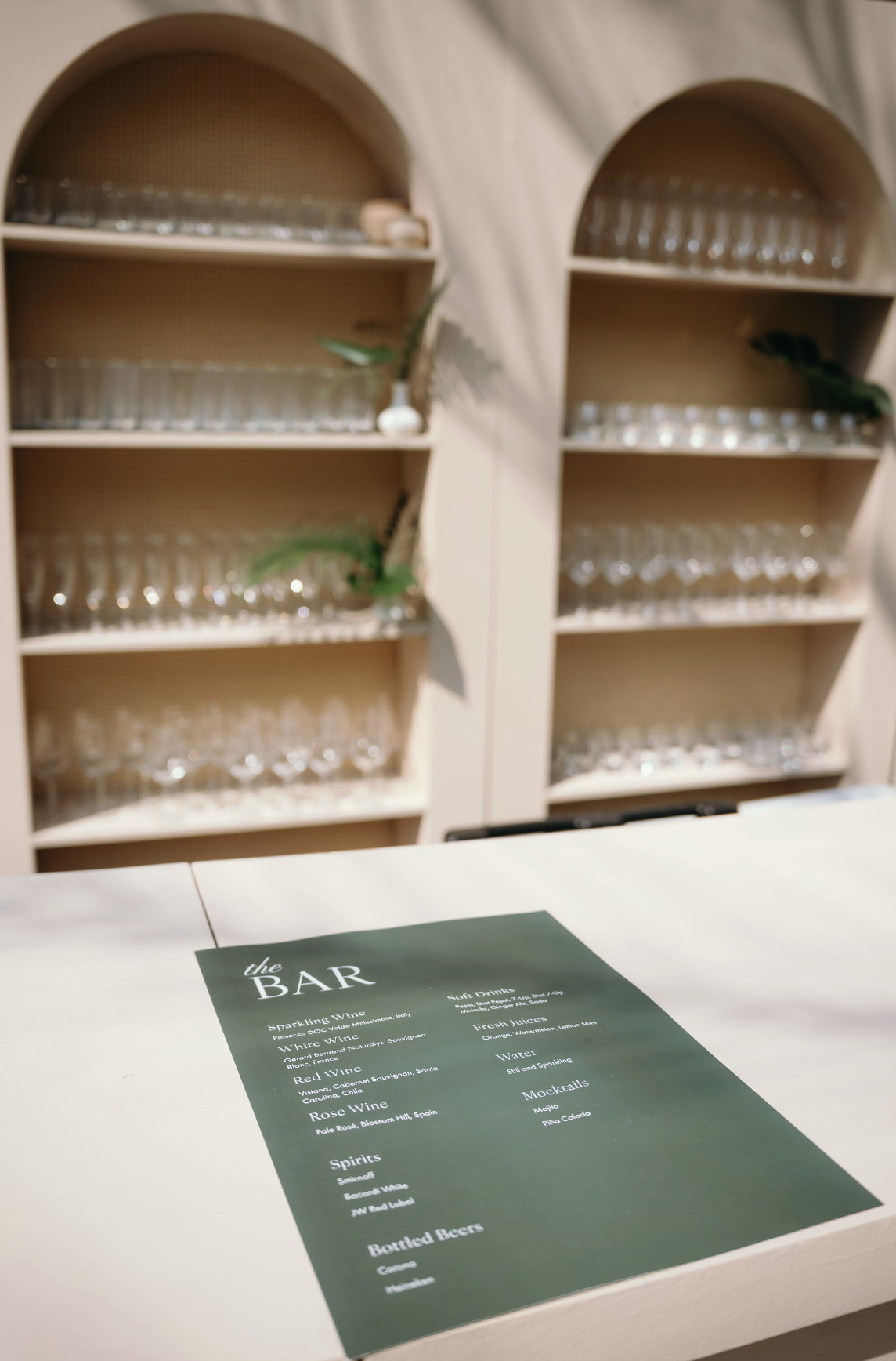 A bar menu on a white table with a dark green background and white text, featuring wine, spirits, and bottled beers, in front of beige shelves with empty glasses.