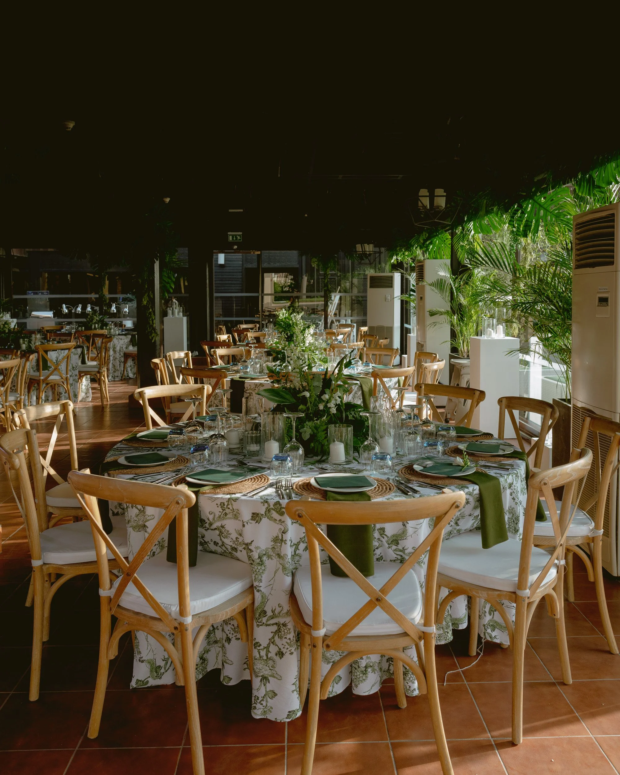 A dining area set up for an event with round tables covered in floral tablecloths, decorated with flowers and candles. Wooden chairs surround the tables, and large windows and lush green plants line the space, creating a bright and inviting atmospher