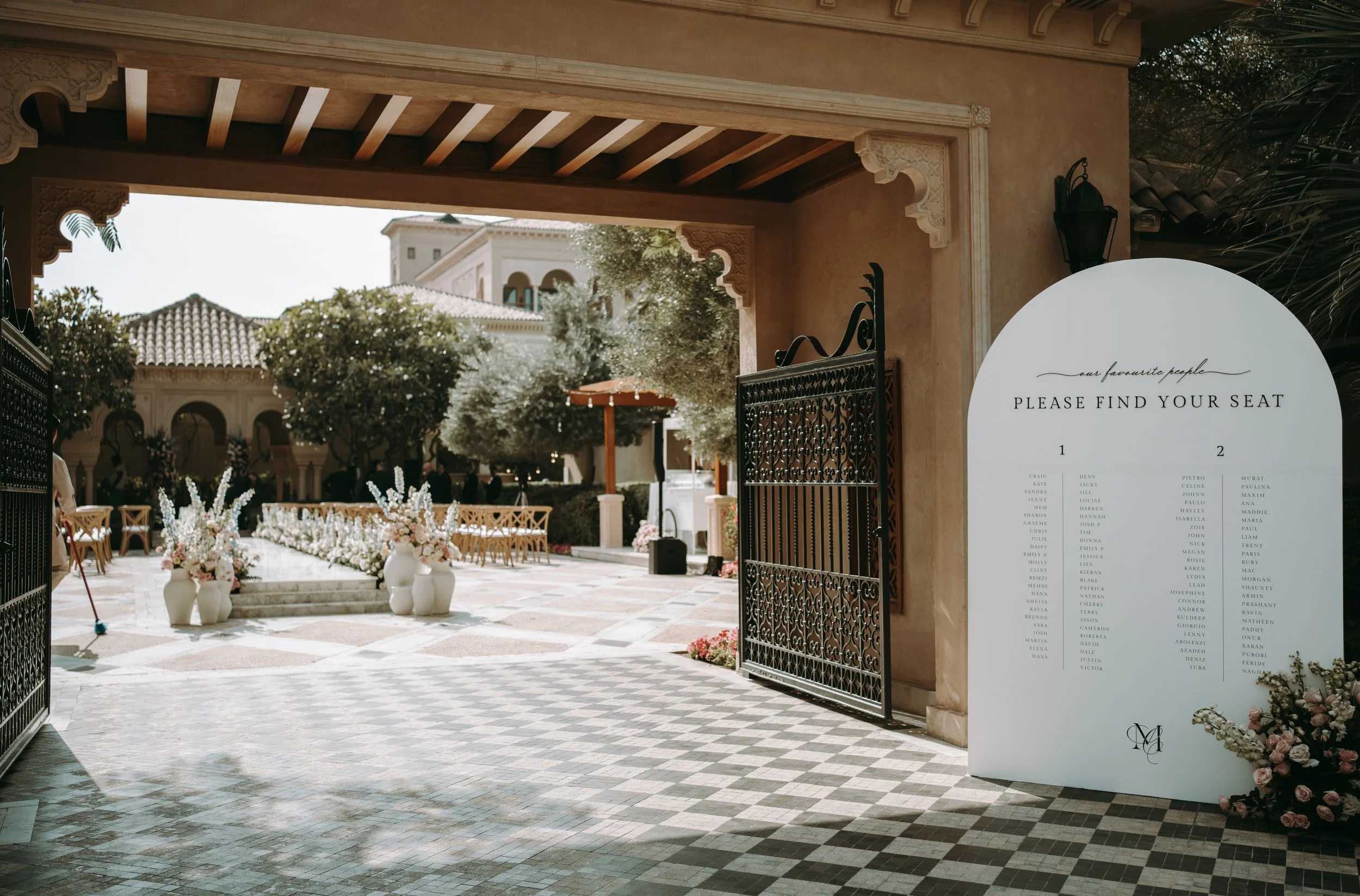 Open gate leading to an outdoor event area decorated with flowers, with chairs and tables arranged for a gathering. A large sign displays a list of names, possibly seating arrangements, near the gate.