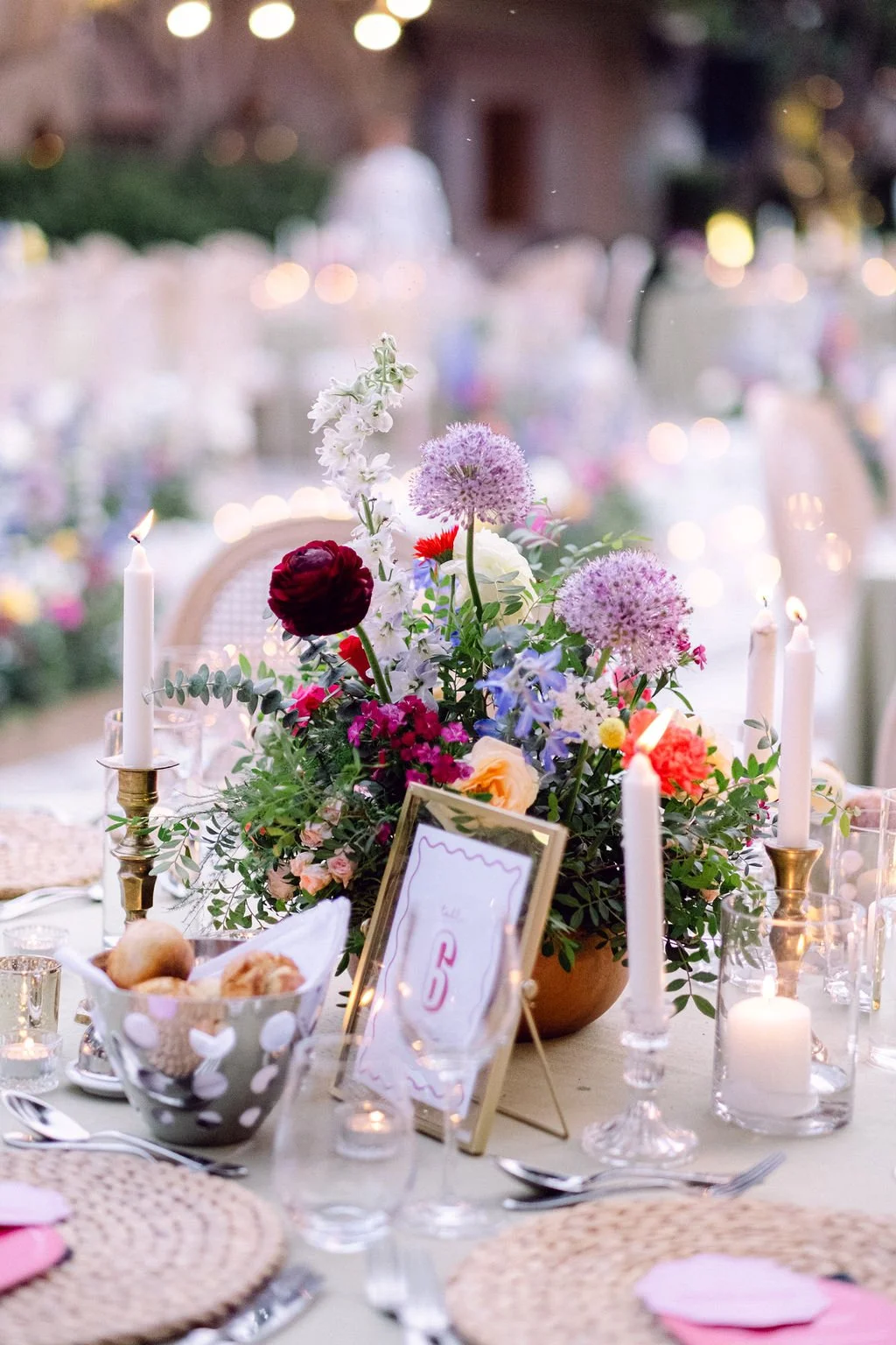 A decorated table at an outdoor event with a floral centerpiece, tall white candles, and framed table number, surrounded by lit candles and place settings with woven placemats.