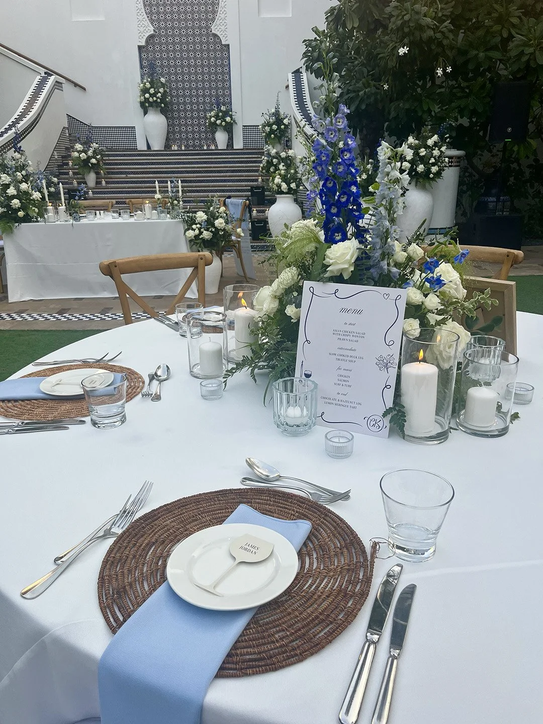 Elegant outdoor wedding reception table with white tablecloth, blue napkin, wicker placemats, glassware, silverware, candles, and a floral centerpiece with white and blue flowers. In the background, a decorated altar with white vases and flowers, and
