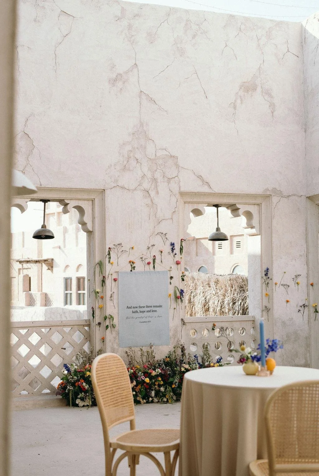 Empty decorated room with round table, chairs, a floral arrangement, and a wall with cracks and decorative cutouts, leading to an outside view of buildings and a field of tall grass.