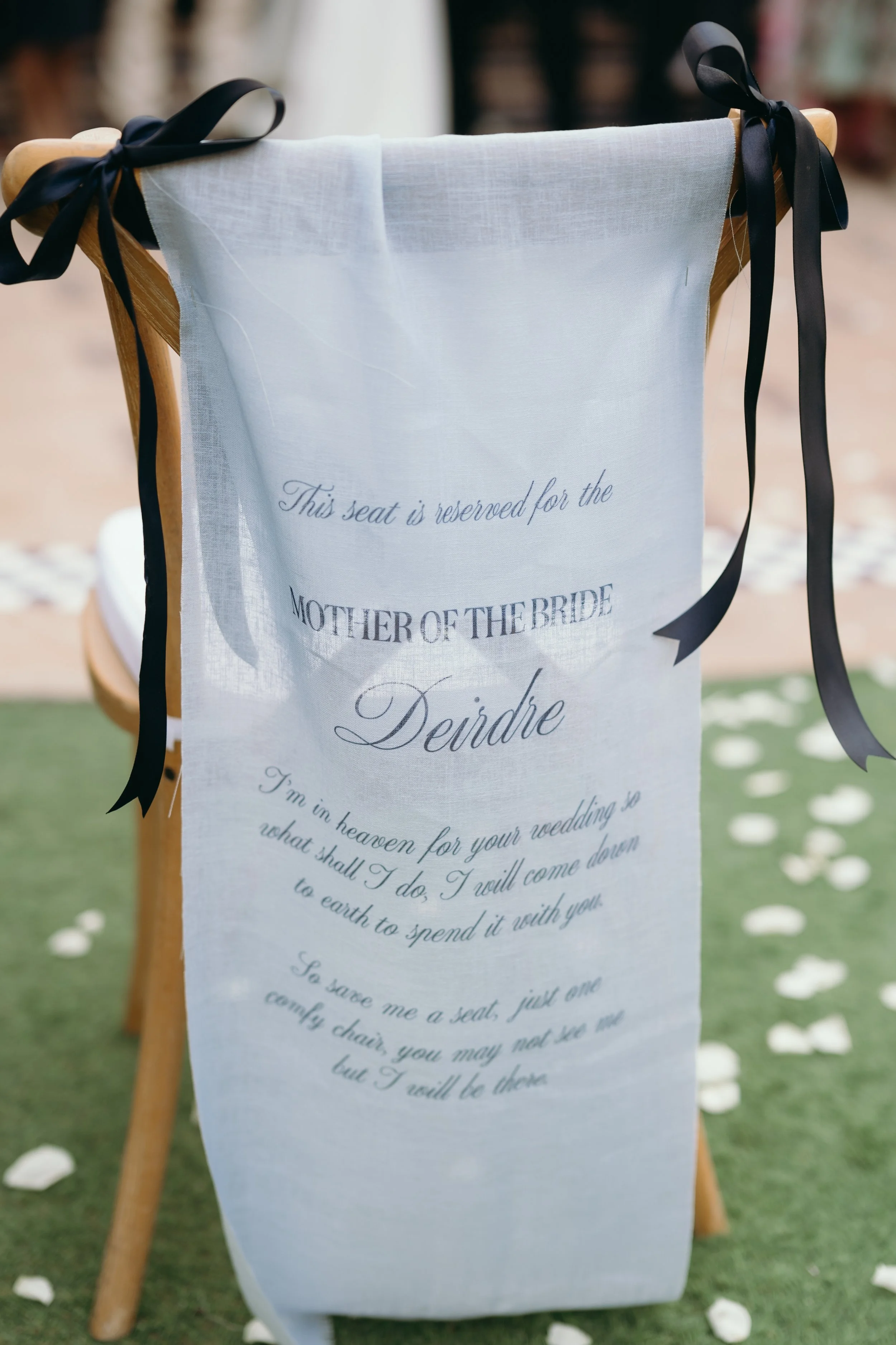 A wooden chair with a white fabric banner draped over it, tied with black ribbons. The banner has words printed on it, including 'This seat is reserved for the Mother of the Bride.' The background shows a blurred indoor setting with a brick wall and 