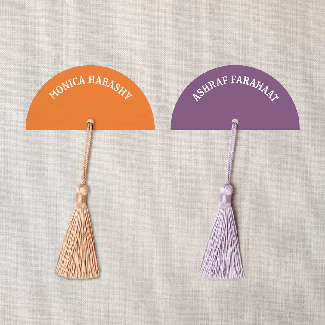 Colorful paper fans with tassels, one orange with name 'Monica Habashy', and one purple with name 'Ashraf Farahat' written on them, pinned to a beige fabric background.