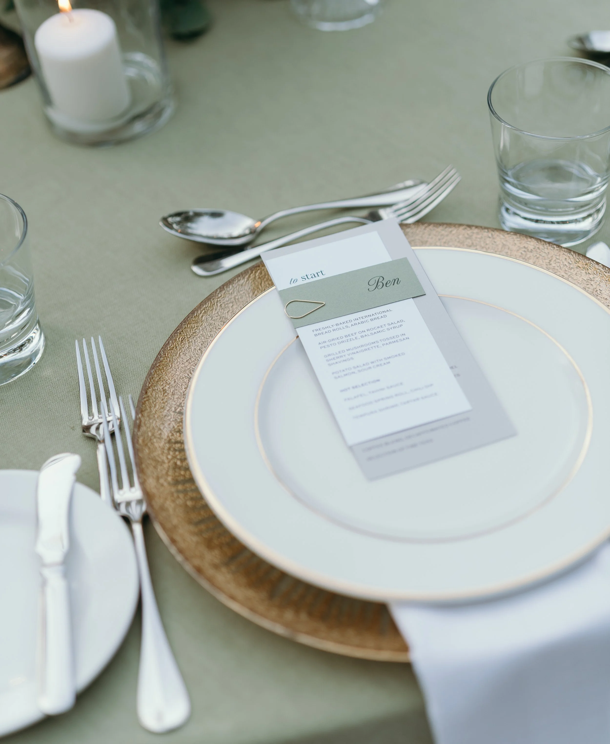 Table setting with a gold-rimmed white plate, a menu card labeled 'Ben', two small forks, a small spoon, and glasses of water on a green tablecloth.