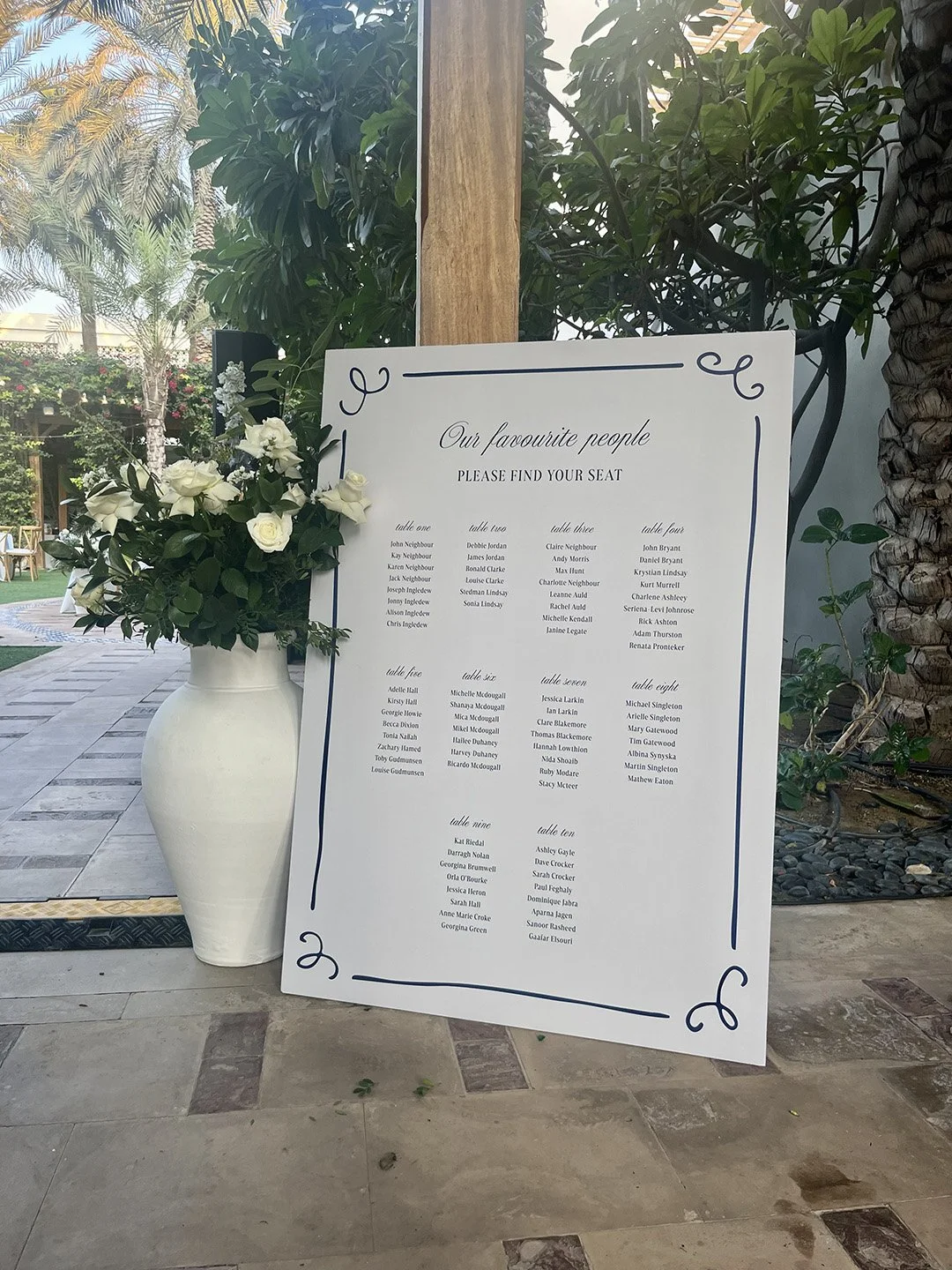 Wedding seating chart with a white and blue design, listing guests' names organized by tables, placed next to a white vase with white roses and greenery outdoors.