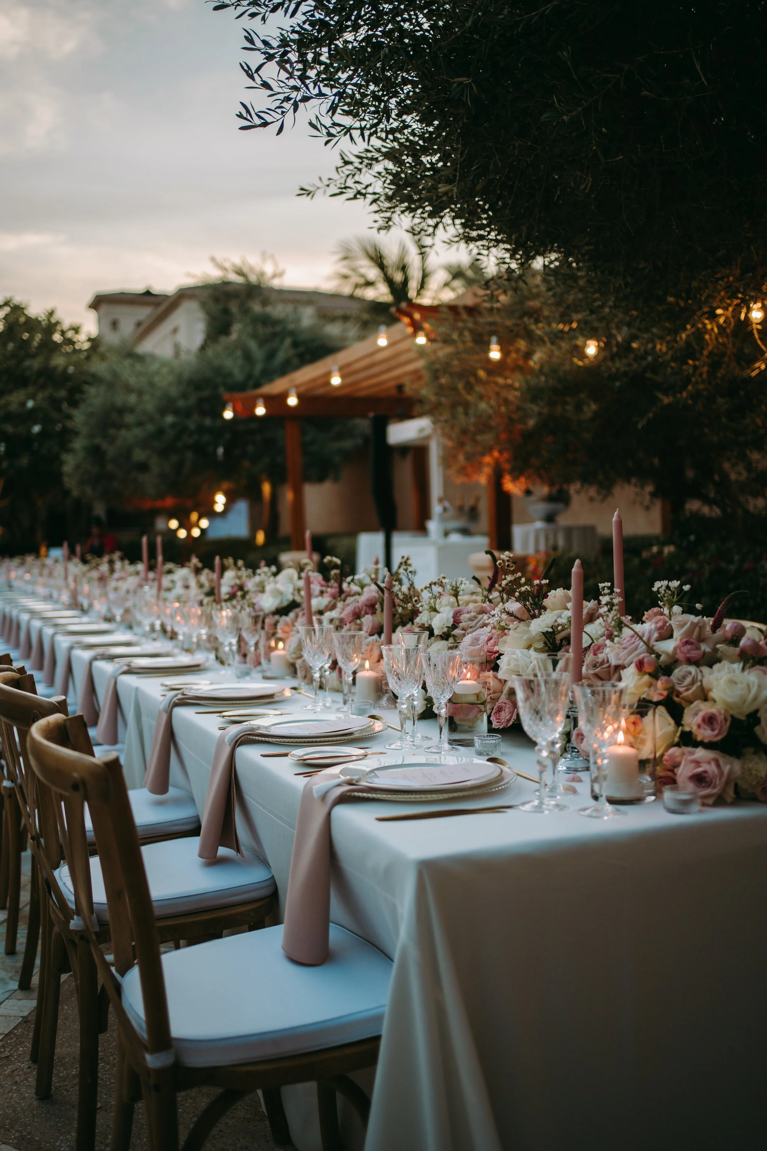 An outdoor dining table decorated with pink and white flowers, candles, and glassware, set for a celebration or event during dusk with string lights and trees in the background.