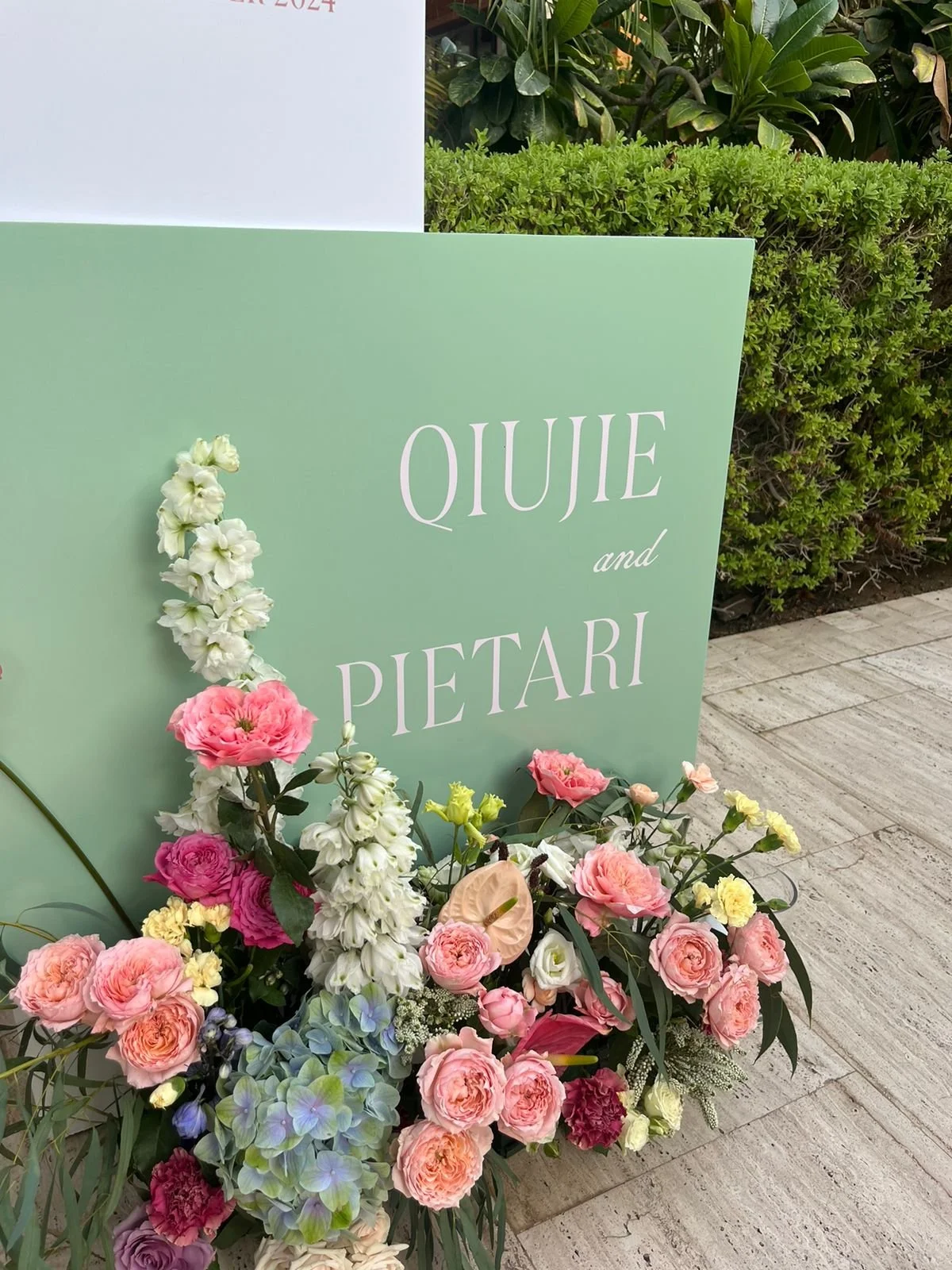 A sign with light green and white colors reading "QIUJIE and PIETARI." Flower arrangements with pink, white, purple, and yellow blossoms are in front of the sign, and green foliage is in the background.