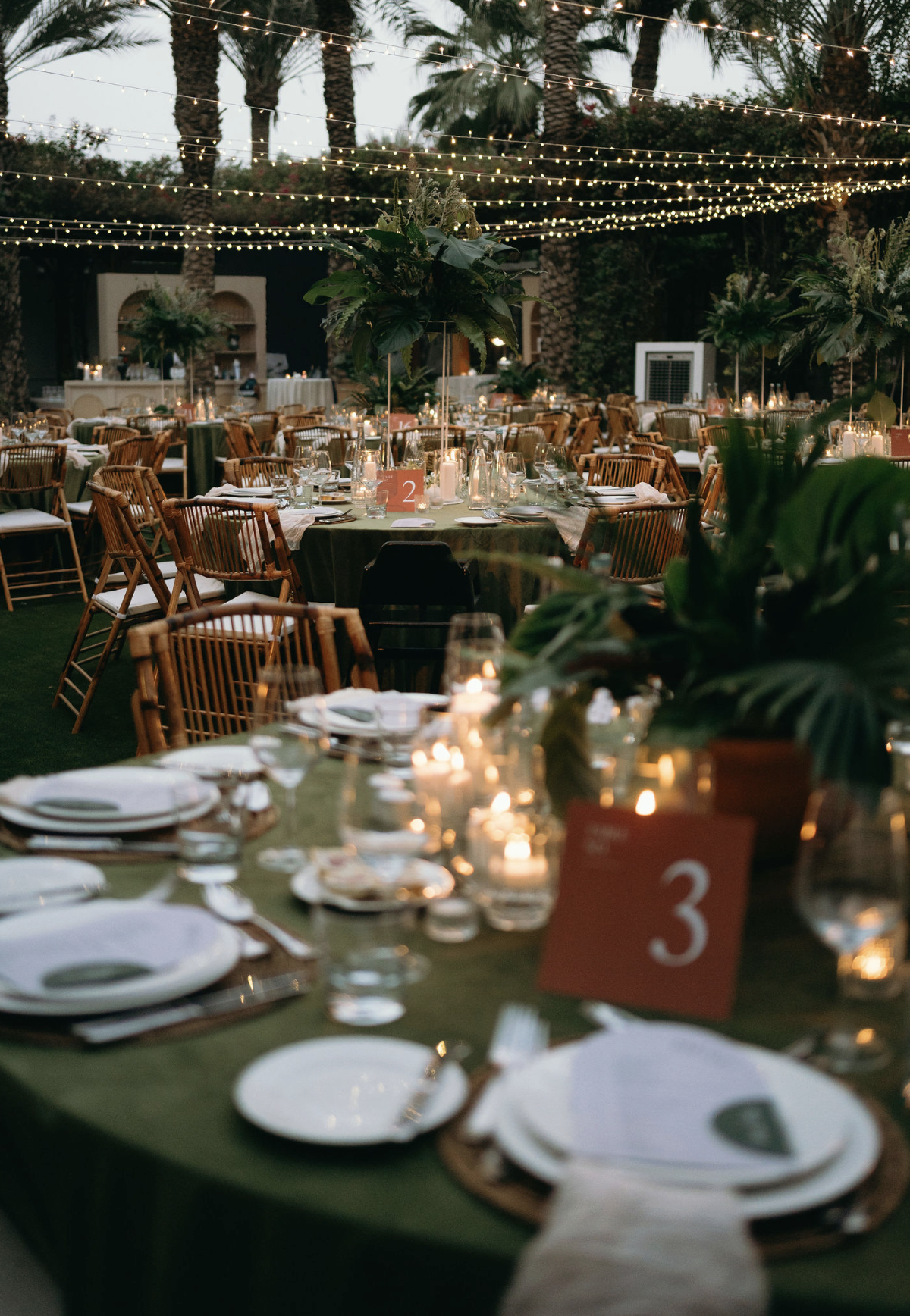 Outdoor evening event with tables decorated with candles, greenery centerpieces, and string lights overhead. Palm trees are visible in the background.