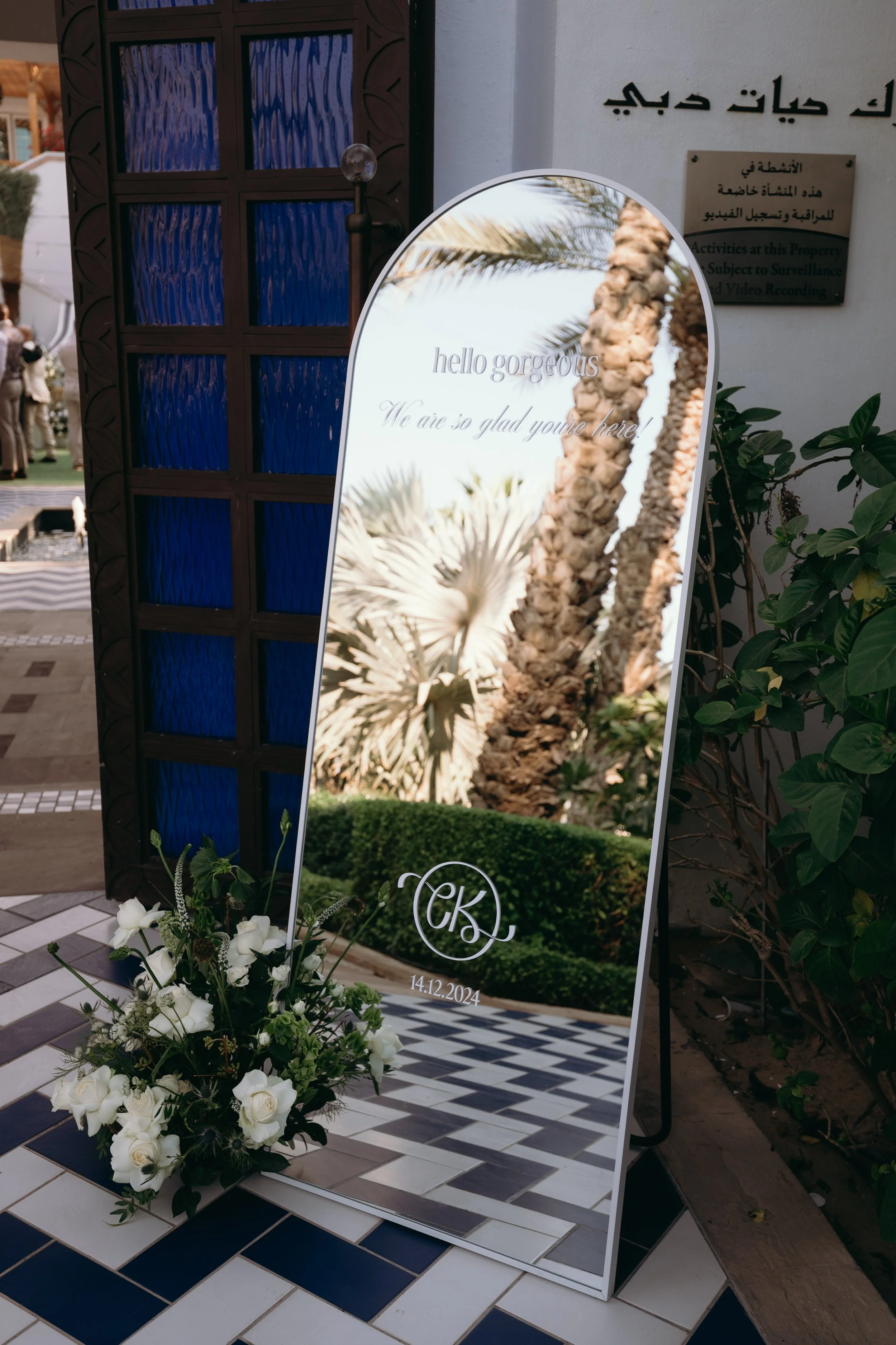 Wedding or event sign with white flowers at the base, displaying the text "hello gorgeous. We are so glad you're here!" and a monogram with the date 14.12.2024, against a backdrop of palm trees and decorative tiles.