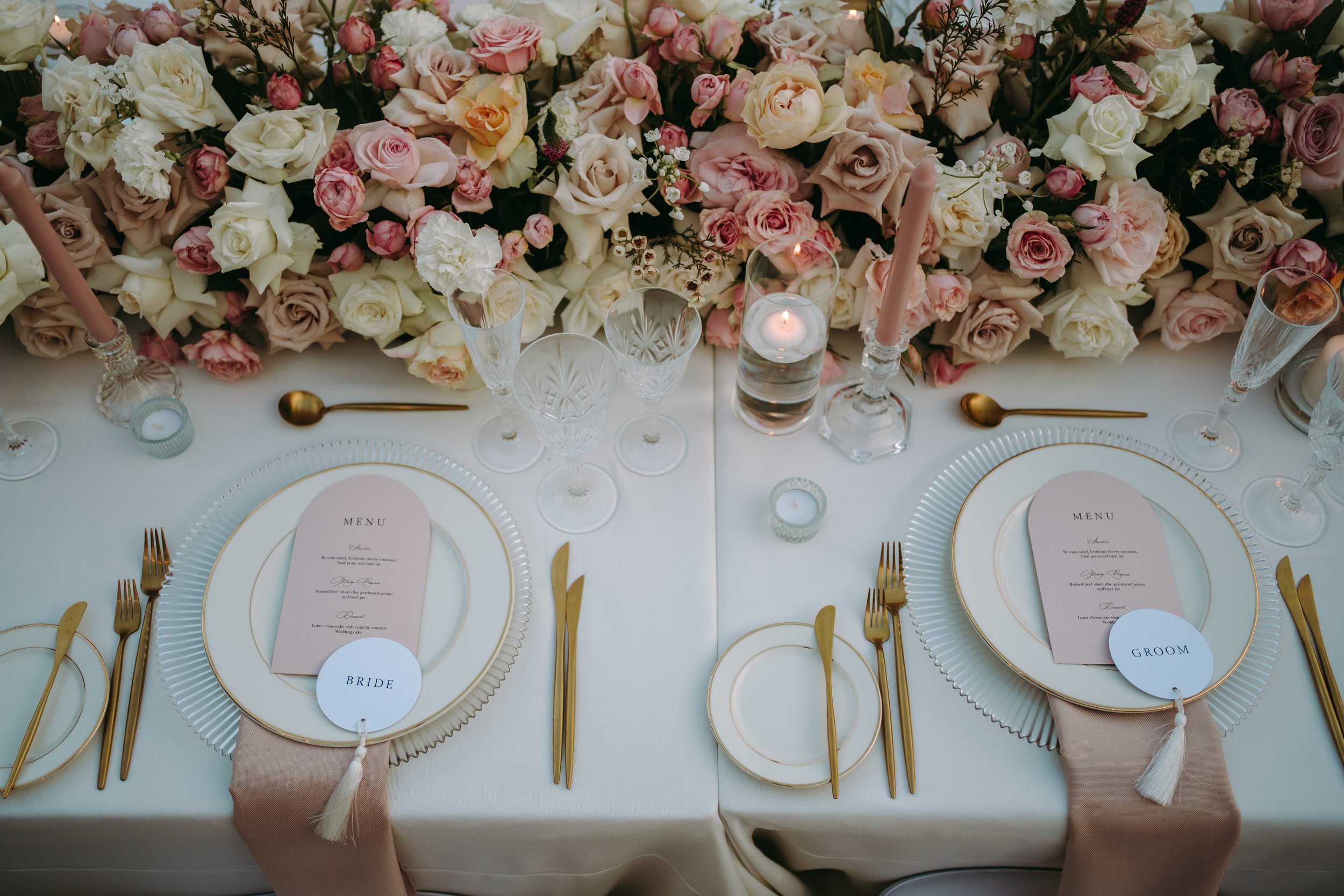 A table setting with a large arrangement of pink and white roses and flowers in the background. The table has place cards labeled 'GROOM', 'BRIDE', and 'GUEST'. There are gold utensils, glassware, a small candle, and pink napkins on the table.