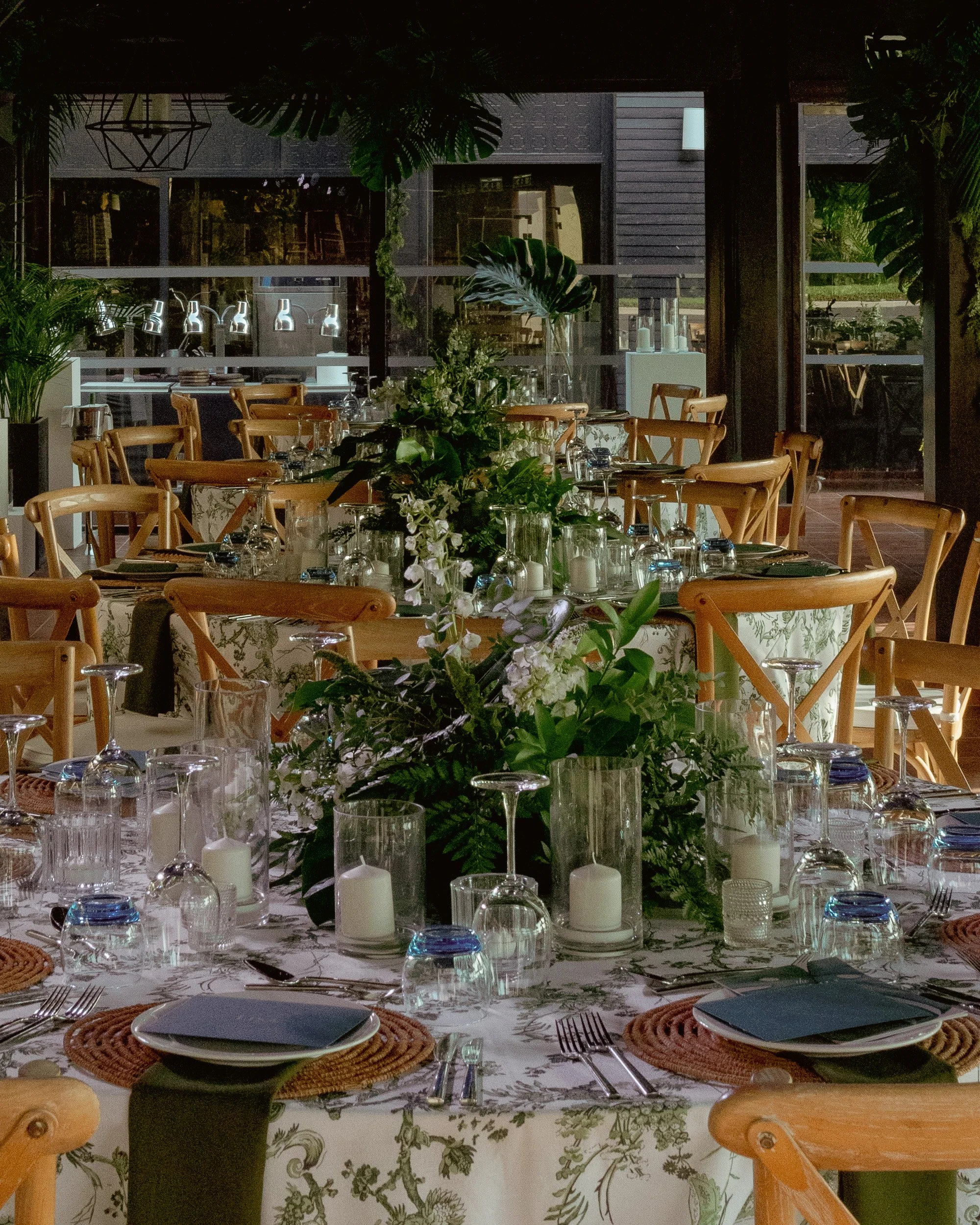 A table set for a formal dinner with floral centerpiece, wine glasses, candles, and place settings, in a room with large windows, greenery, and modern decor.