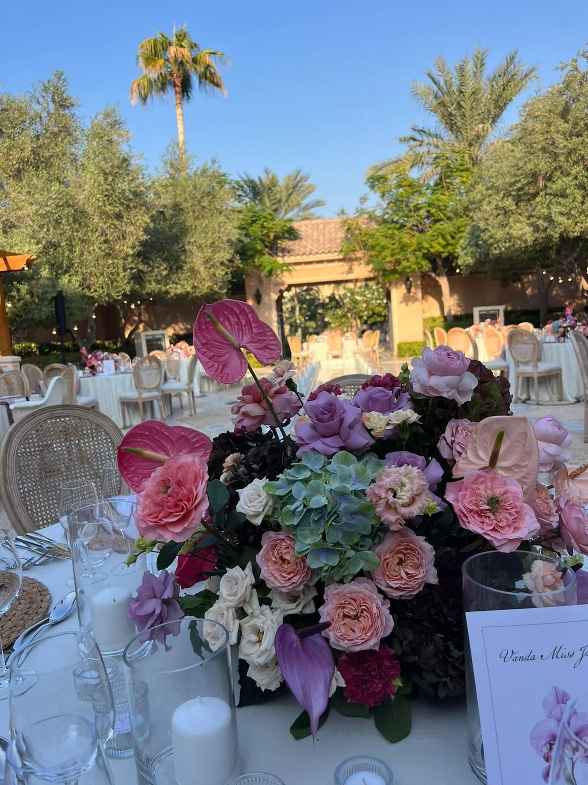 A decorated outdoor event space with a floral centerpiece featuring pink, purple, and white flowers, set on a table with glassware and candles. In the background, there are round tables with chairs, surrounded by trees and a building with a tiled roo