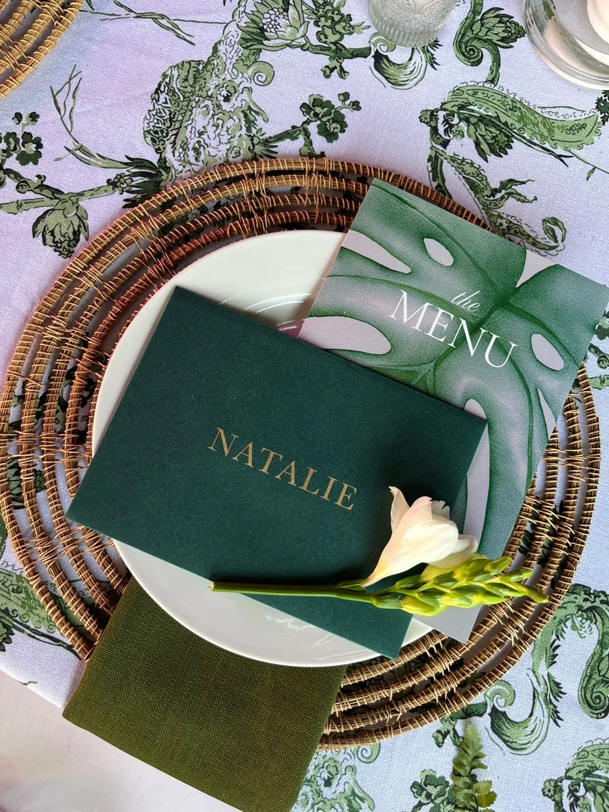 A table setting featuring a white plate on a woven placemat, with a green booklet labeled 'NATALIE,' a menu card with a tropical leaf design, a white flower, and a glass of water.