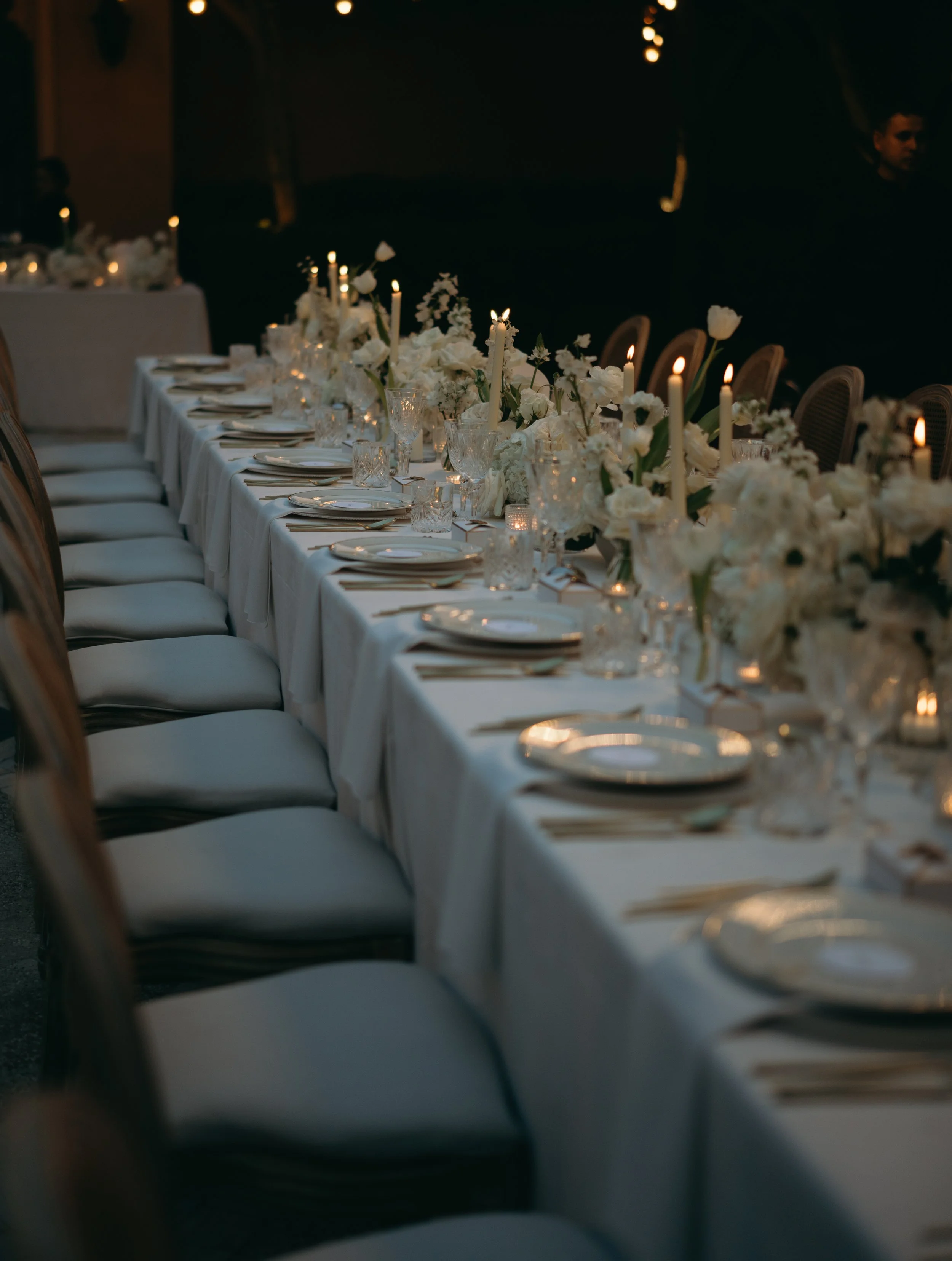 A long dining table decorated with white flowers, candles, and tableware, set up for a formal event, with chairs on either side, in a darkened room with soft lighting.