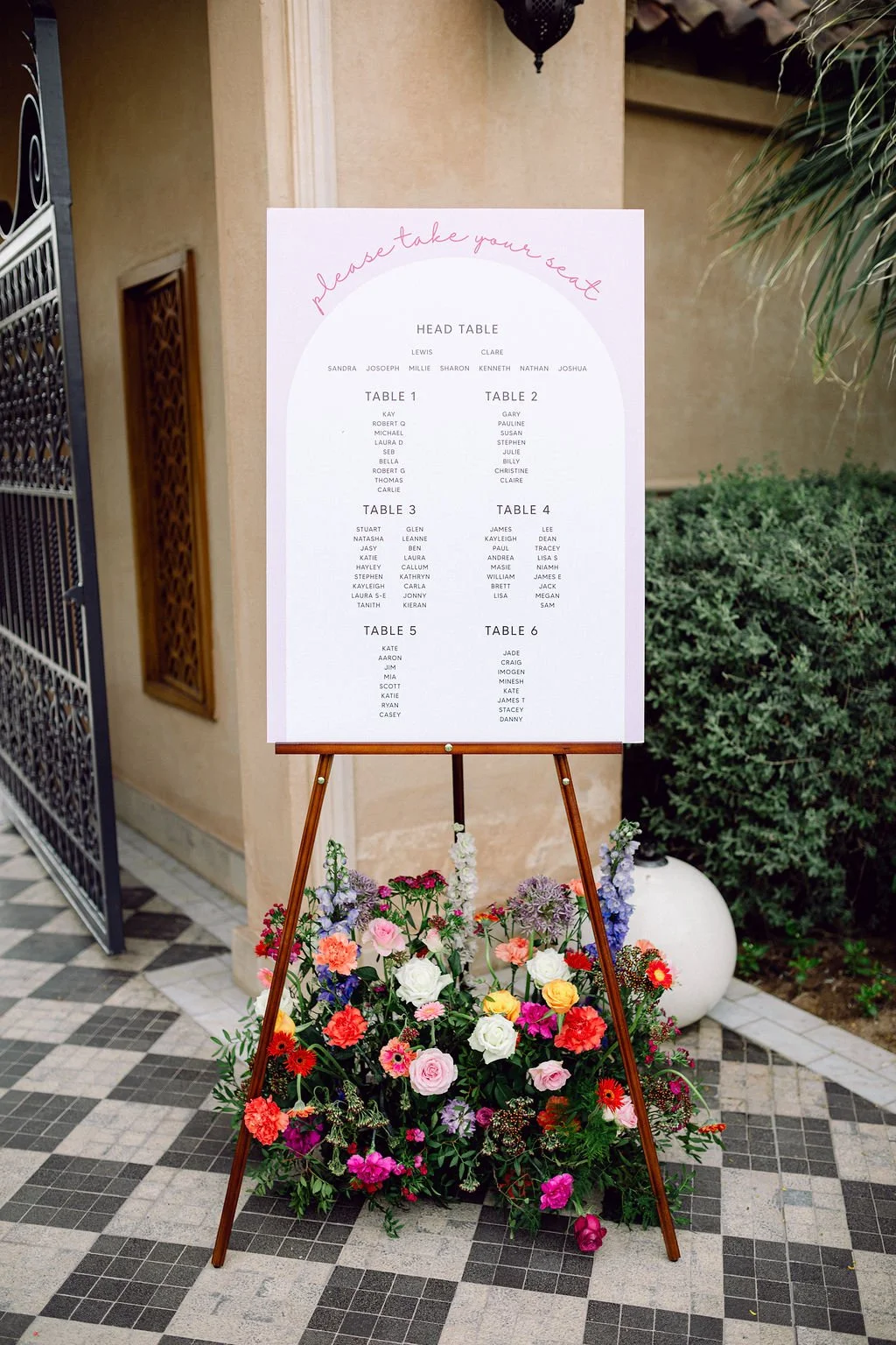 Wedding seating chart on an easel surrounded by colorful flowers, with a gate and greenery in the background.