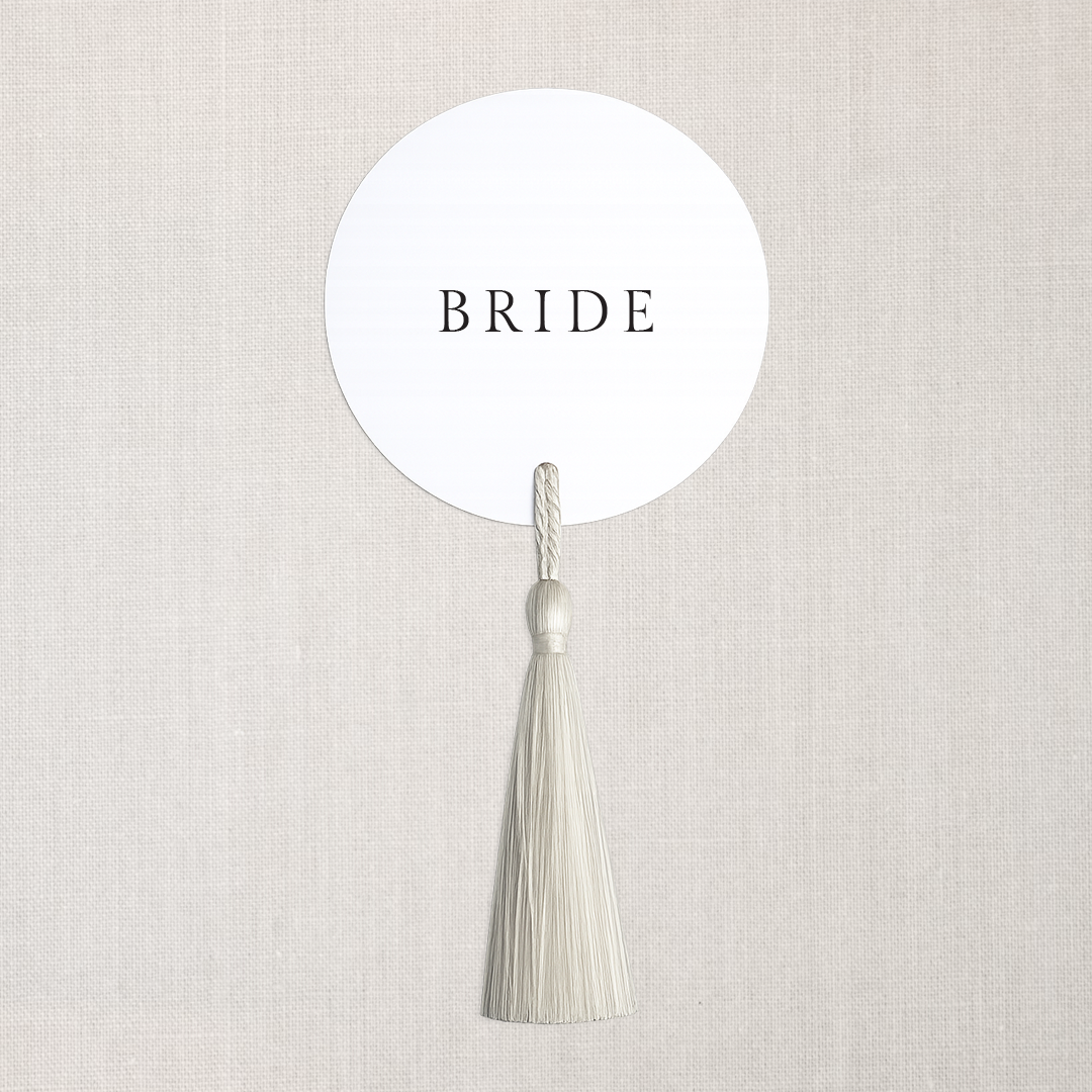Round white sign with the word 'BRIDE' in black letters, hanging from a white tassel attached to a white string against a neutral background.