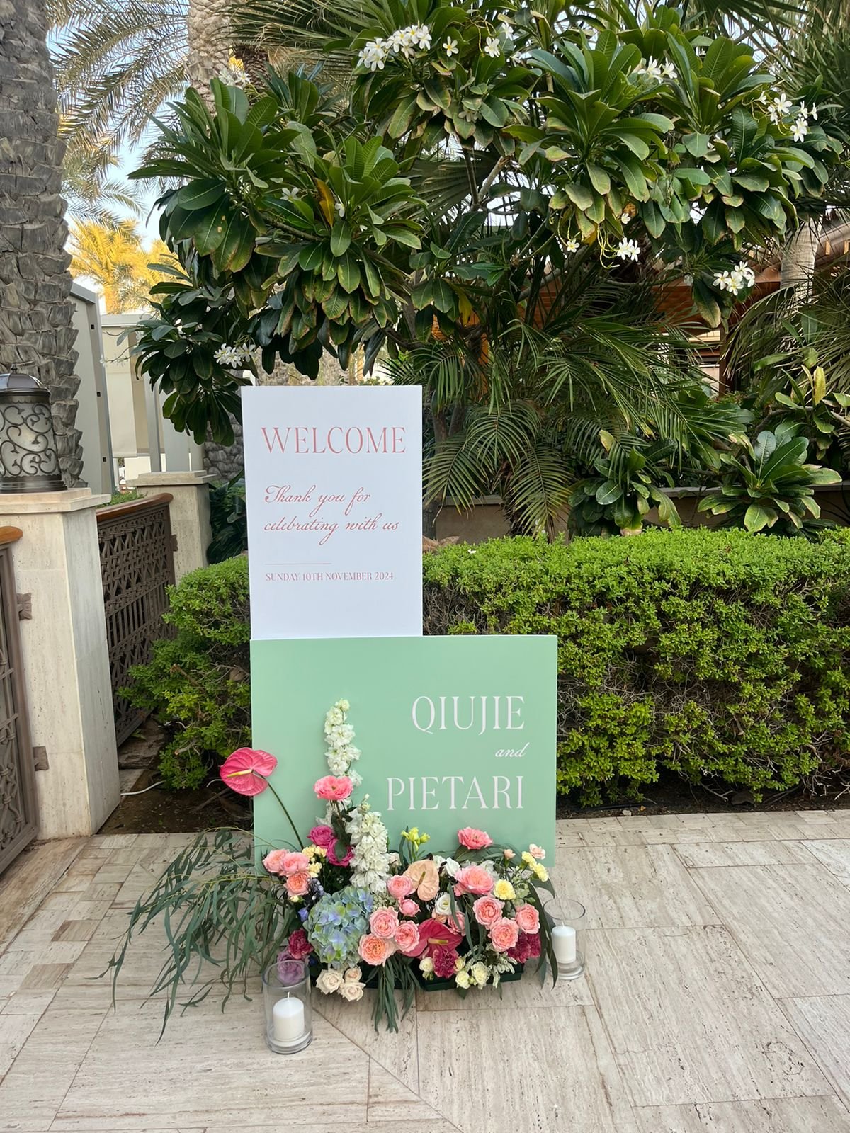 Wedding welcome sign with flower arrangement outside a building. The sign reads 'WELCOME Thank you for celebrating with us SUNDAY 10TH NOVEMBER 2024' and below it another sign says 'QIJIJE and PIETARI' with a colorful flower arrangement at the base, 