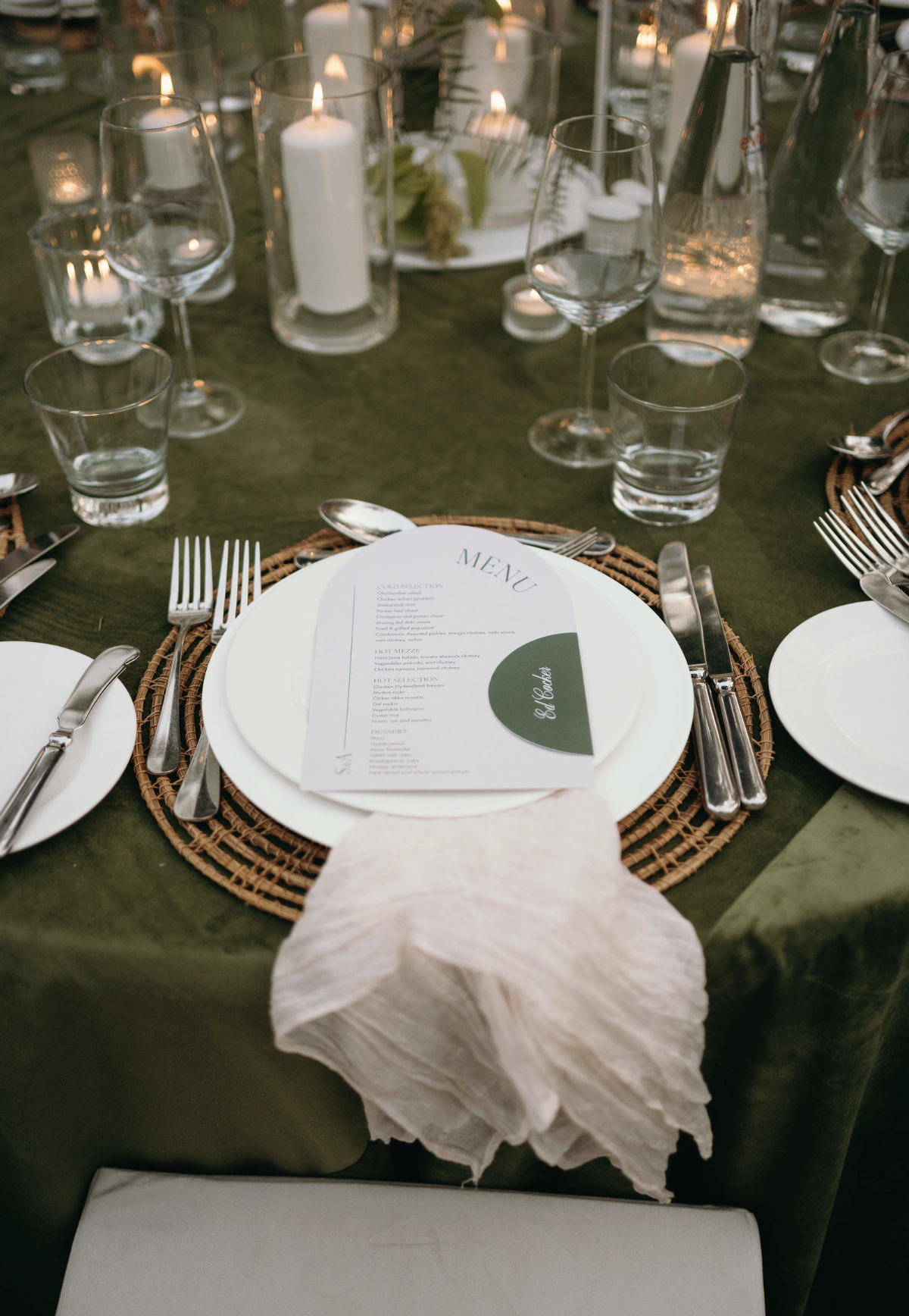 A neatly set dining table with a dark green tablecloth, glassware, silverware, and a menu on a white plate. The table is decorated with candles in glass holders and a centerpiece with greenery. A white cloth napkin is draped over a chair at the front