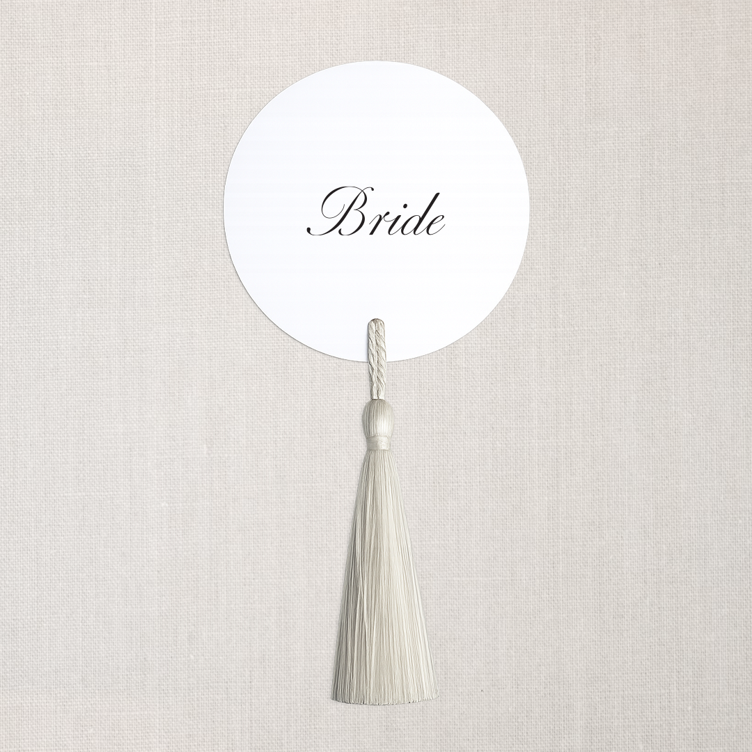 A white circular sign with the word 'Bride' in elegant black script, hanging from a white tassel against a plain, light-colored background.