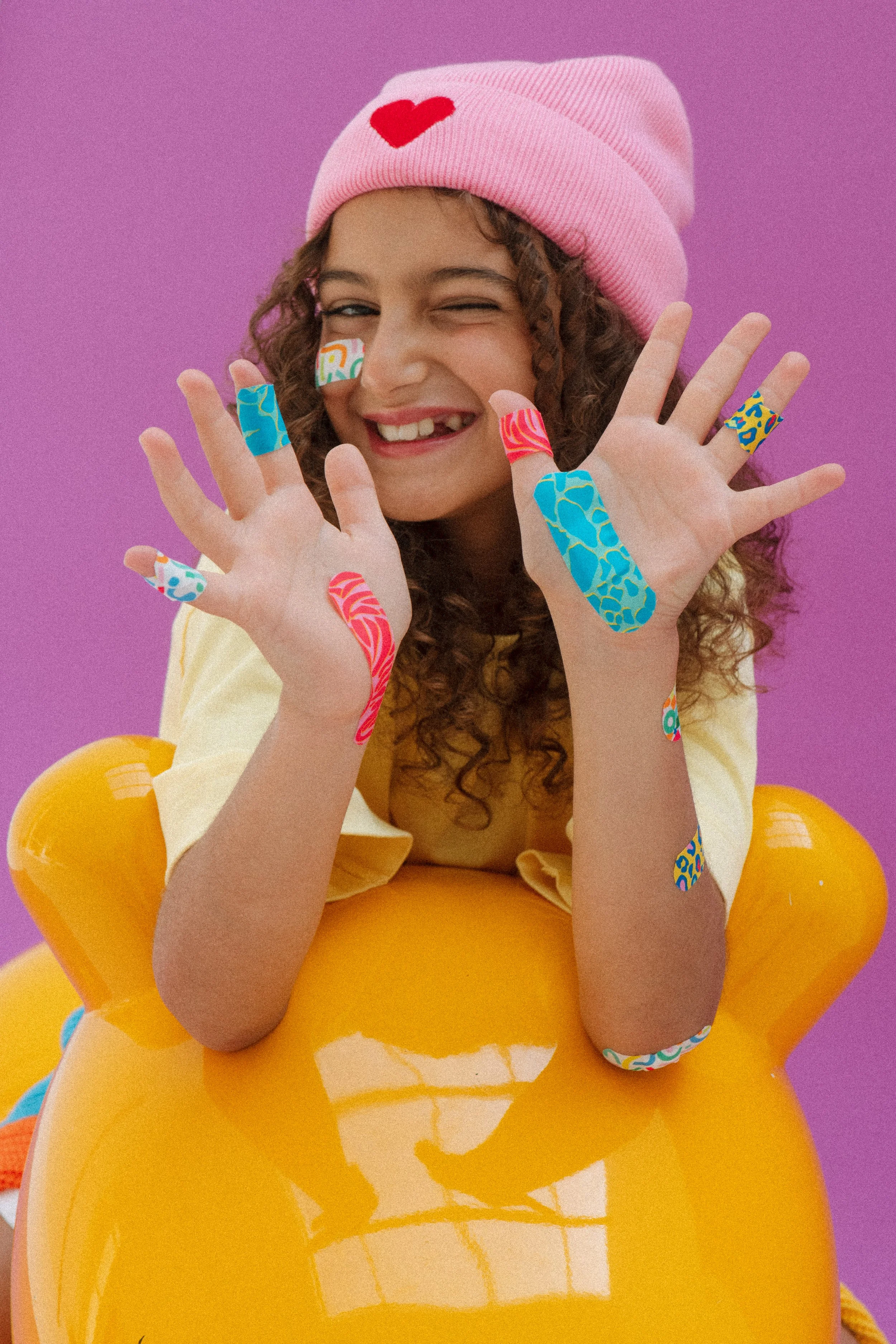 A young woman with curly hair smiling and winking, wearing a pink beanie with a red heart, a yellow top, and yellow inflatable armbands. She has colorful tape on her fingers and cheeks against a purple background.