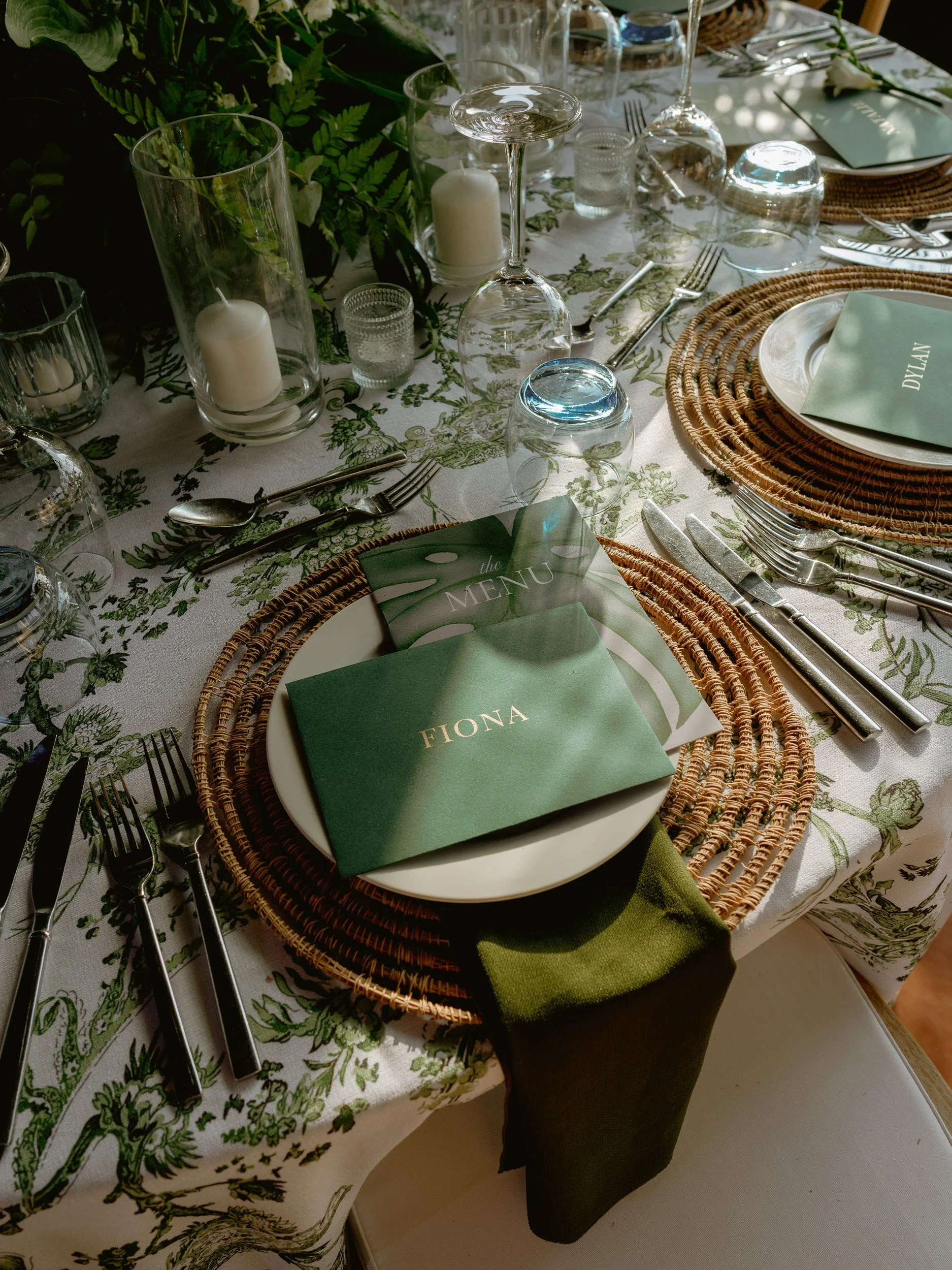 A formal dining table set with plates, silverware, wine glasses, candles, greenery, and green place cards with the name Fiona on one of them, arranged with woven chargers and napkins.