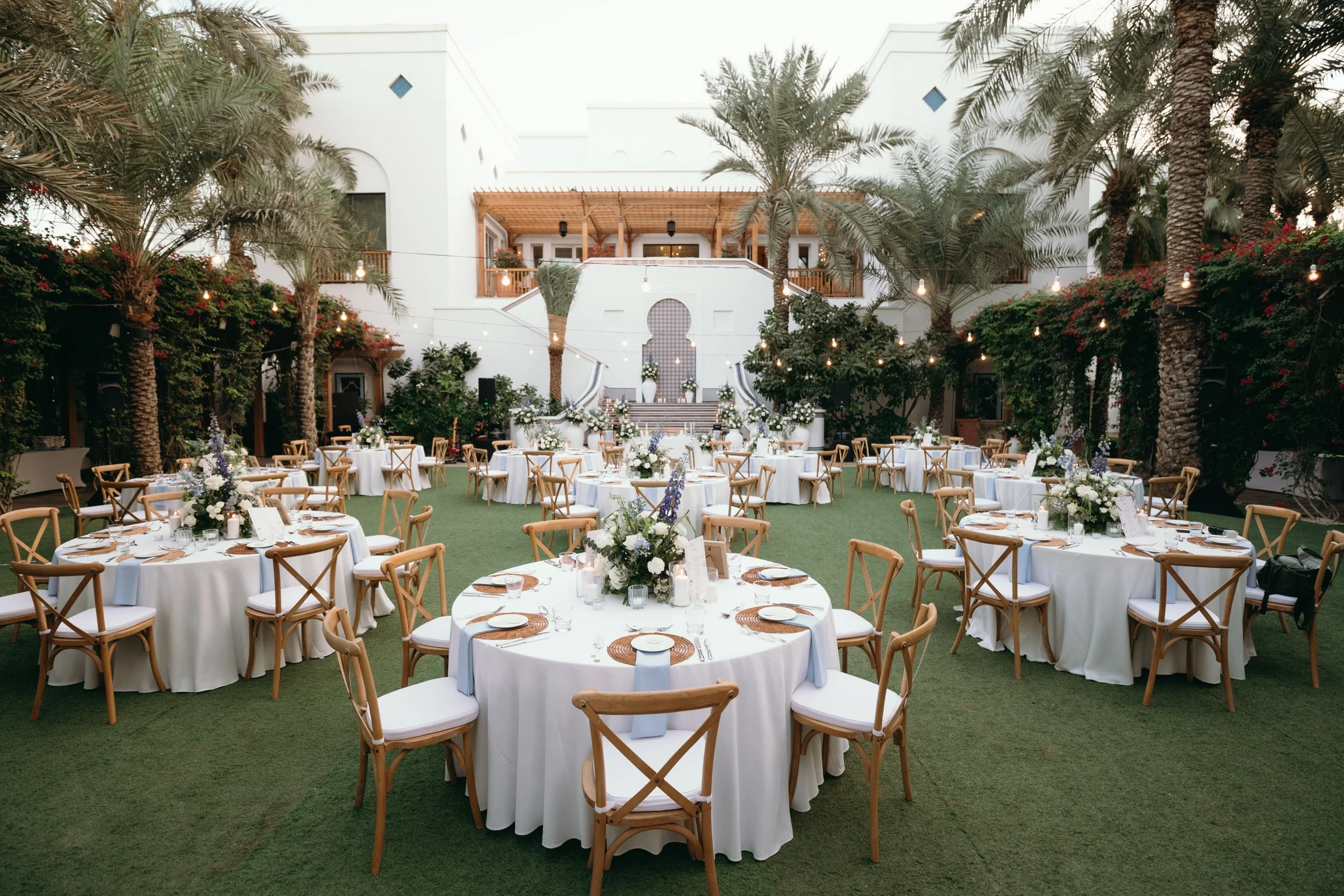 Outdoor event setup with round tables draped in white tablecloths, decorated with floral centerpieces and candles, surrounded by wooden chairs, set on green artificial grass with palm trees and a white building in the background, and string lights ha
