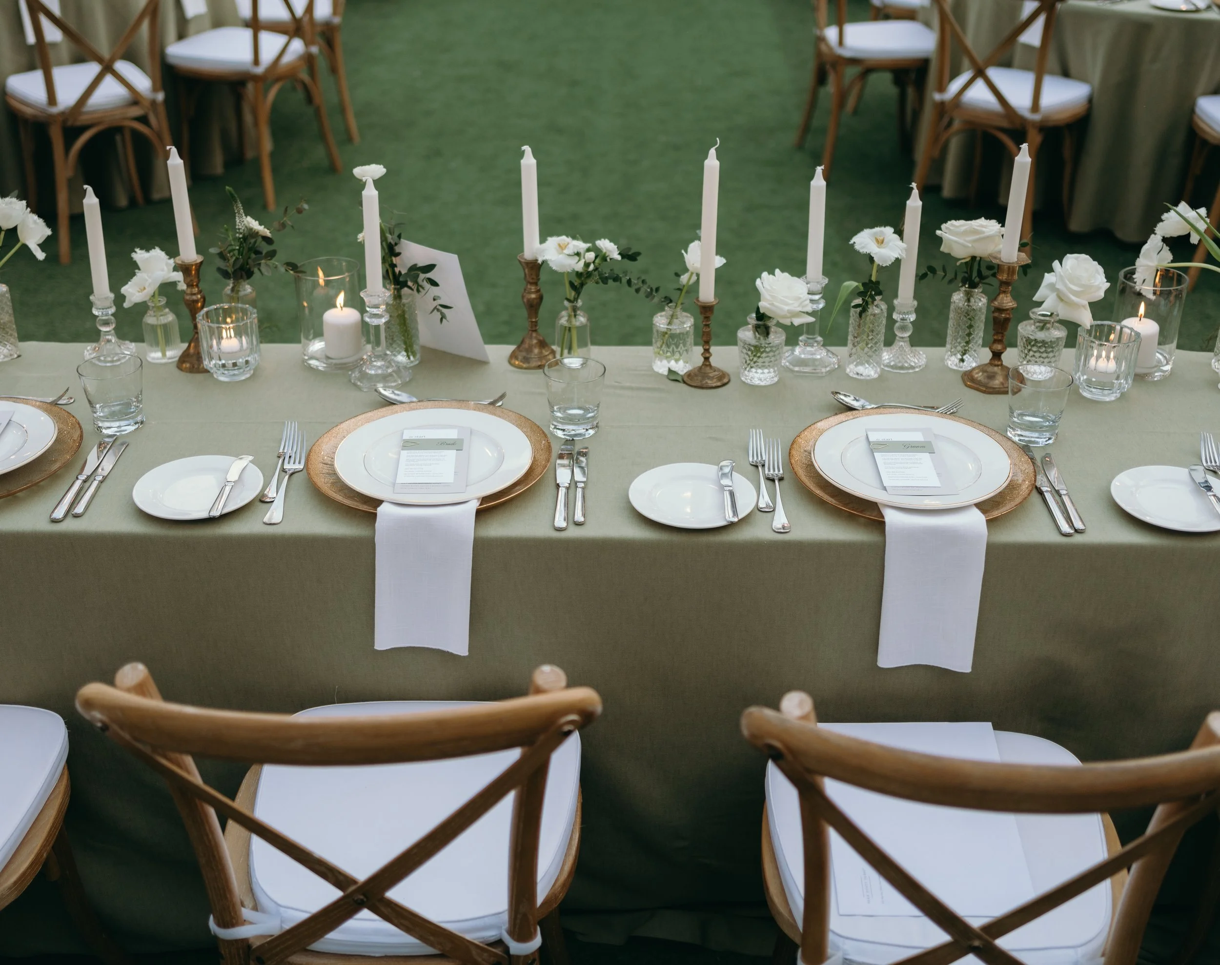 Wedding reception table with floral centerpieces, white candles, and place settings, with green grass and gold chairs in the background.