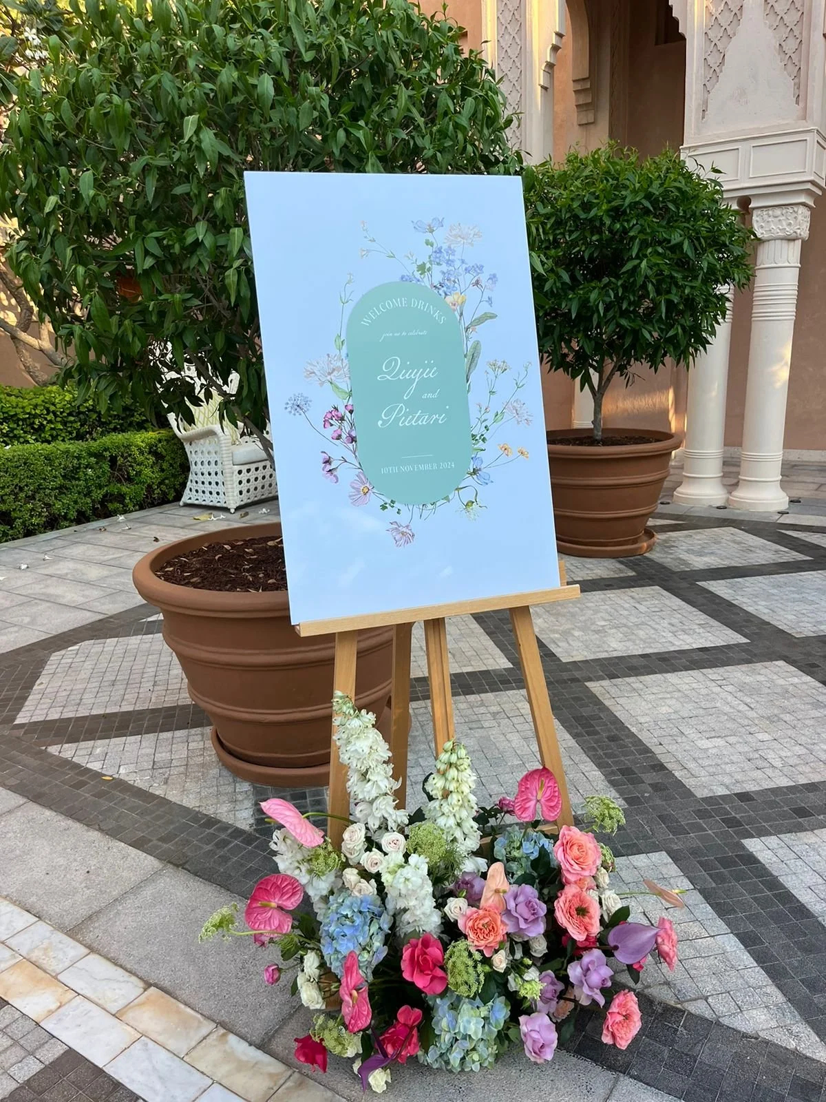 Wedding welcome sign on an easel with floral decoration at the base, placed in an outdoor setting with potted trees and patterned tiled pavement. 