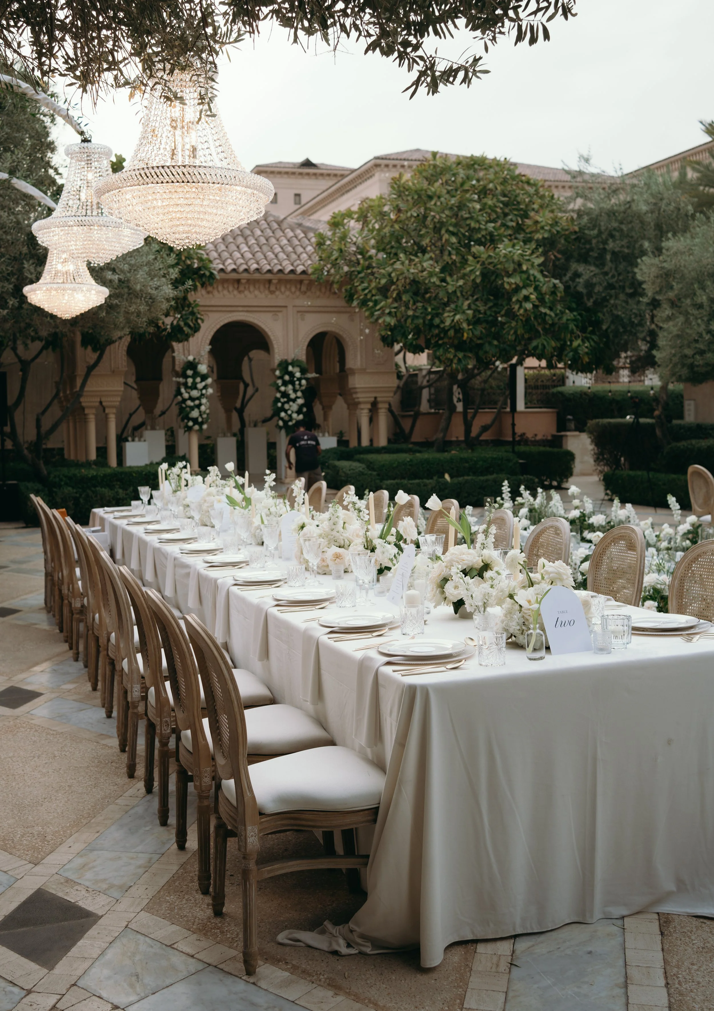 Elegant outdoor wedding reception table with white linens and floral centerpieces, surrounded by chairs, under chandeliers, with a grand building and lush trees in the background.