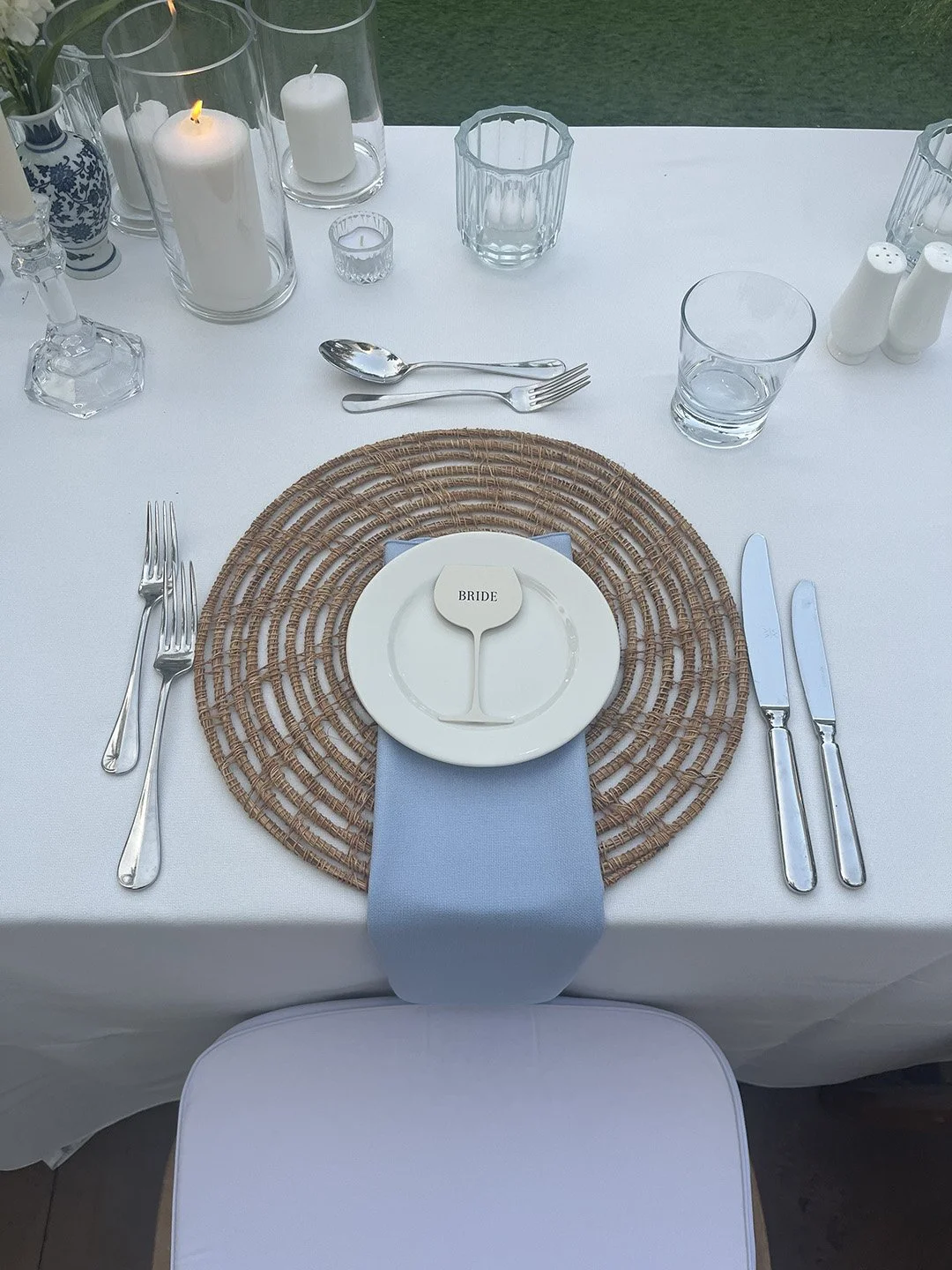 Elegant table setting with a woven placemat, white dinnerware, silverware, and candles, with a officiant or bride place card on a small plate.