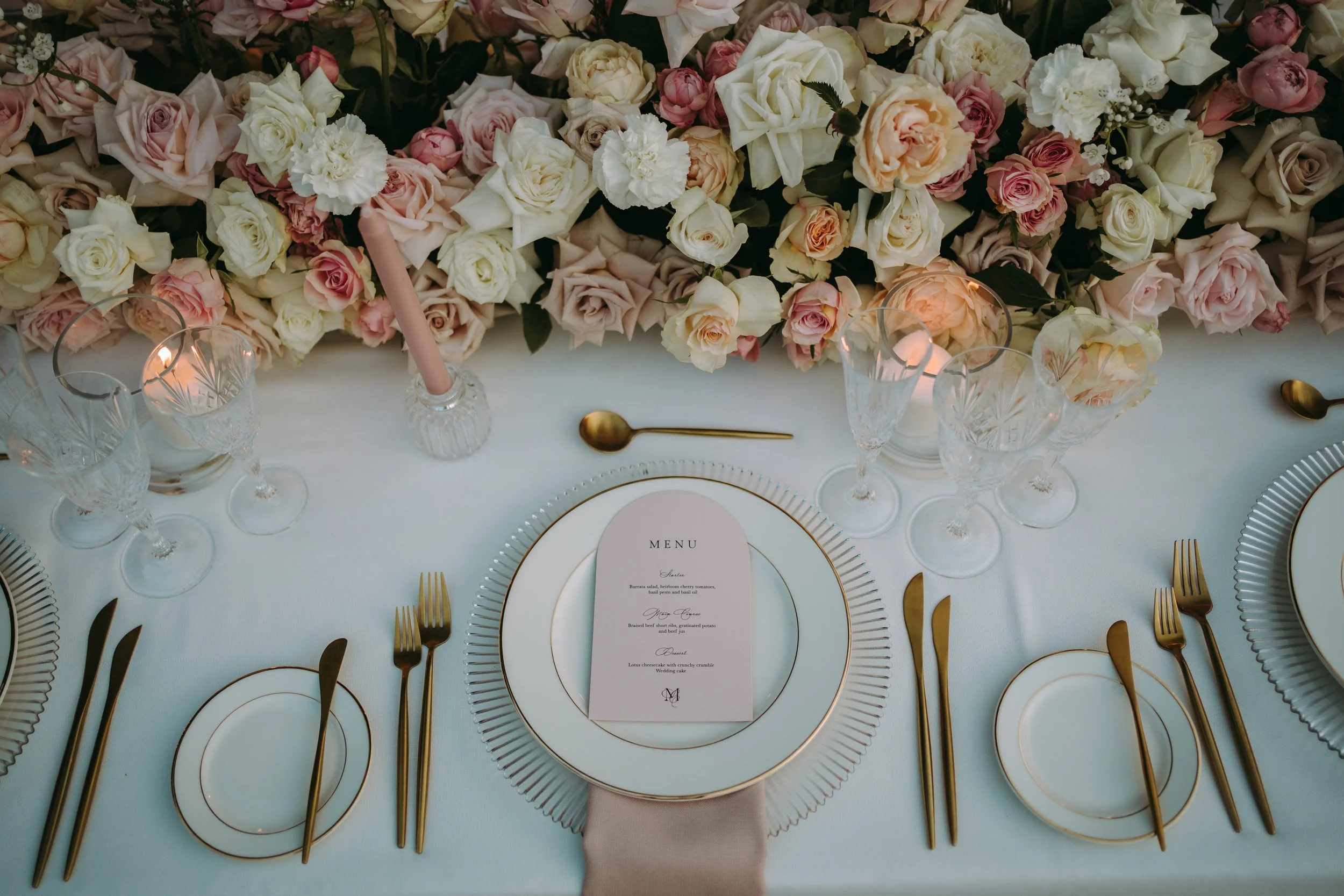 Elegant dining table decorated with a large floral centerpiece of pink and white roses, surrounded by crystal glasses, gold cutlery, and fine china. A wedding menu card is placed in the center of the table.