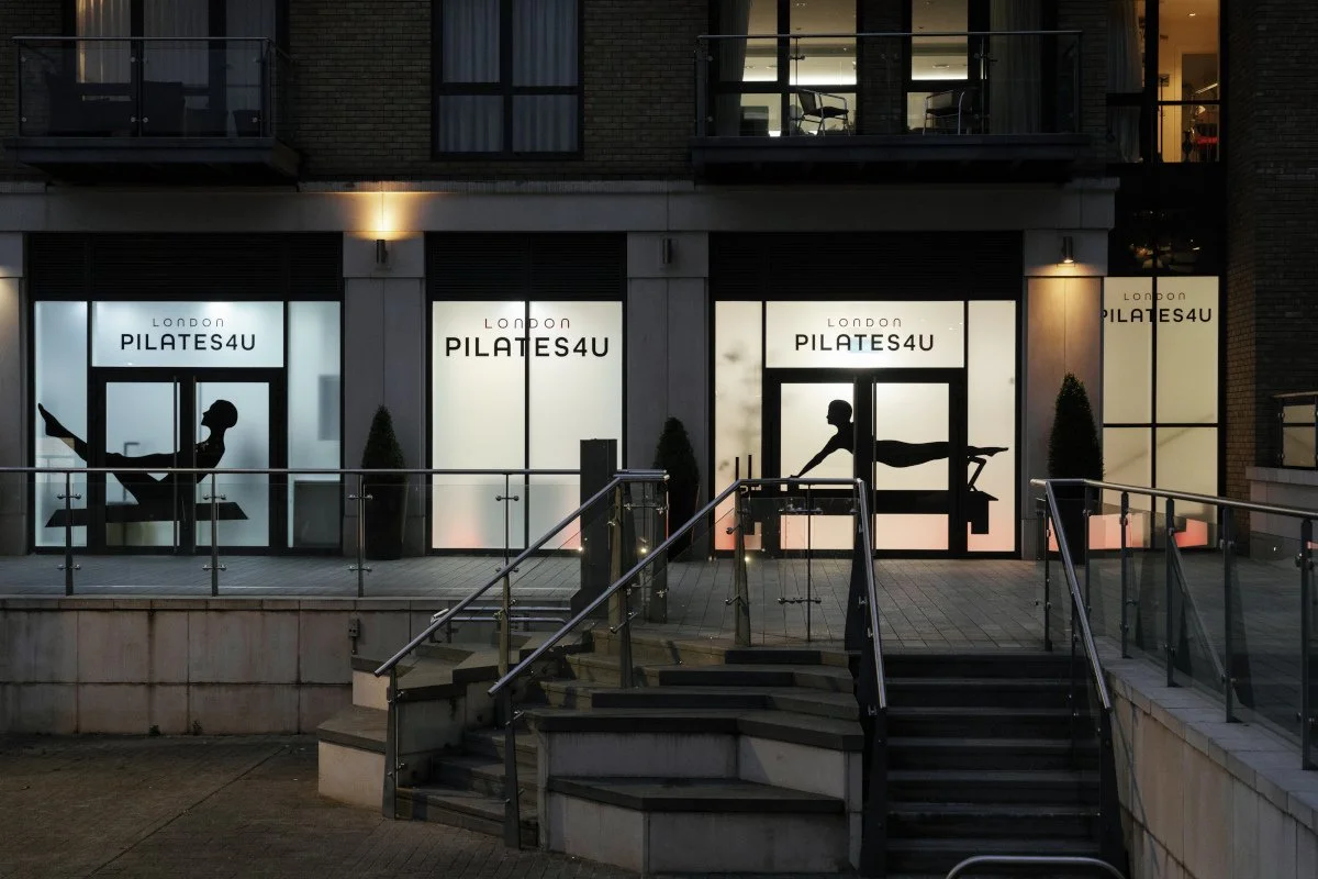 Storefront with four illuminated windows displaying silhouettes of people practicing Pilates, with the signs reading 'London PILATES4U'.